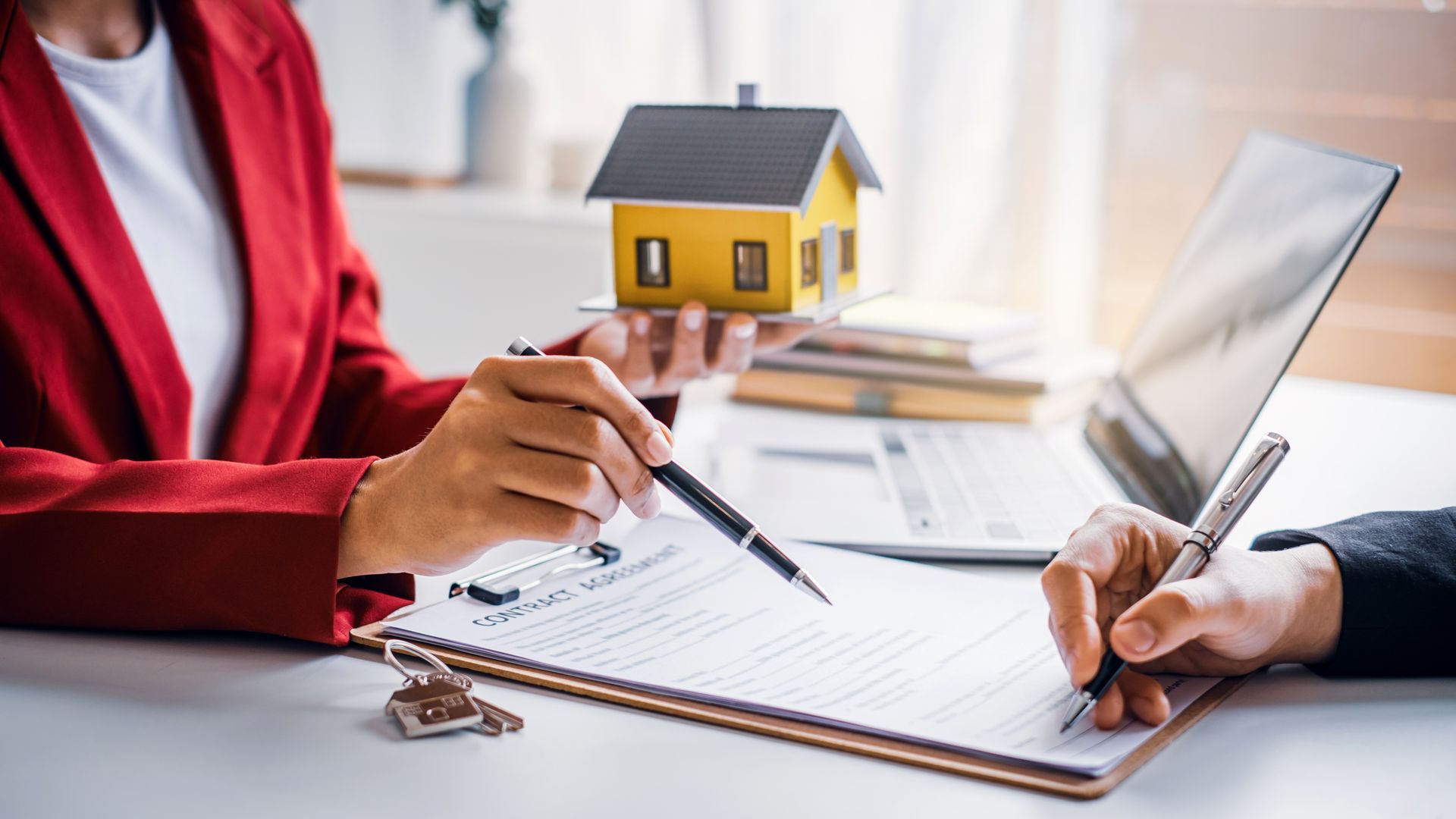 Woman in red jacket pointing at paperwork as another person signs, house model present.