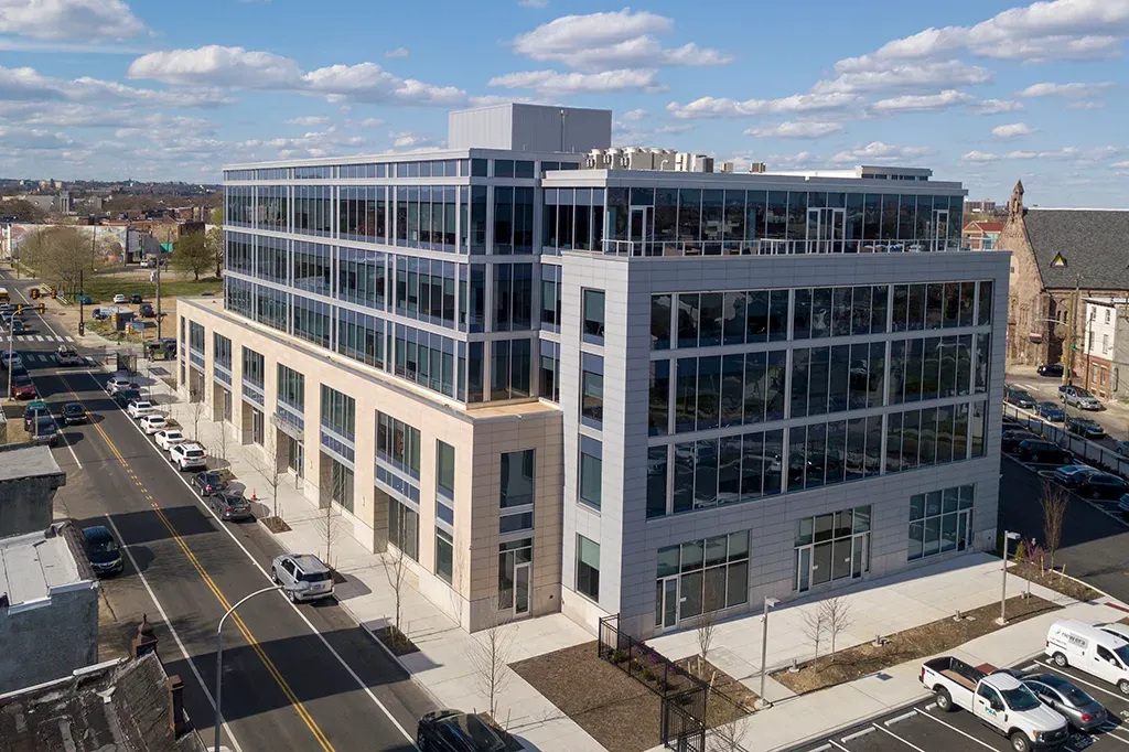 Modern multi-story office building with many windows, next to a street with cars and parking.