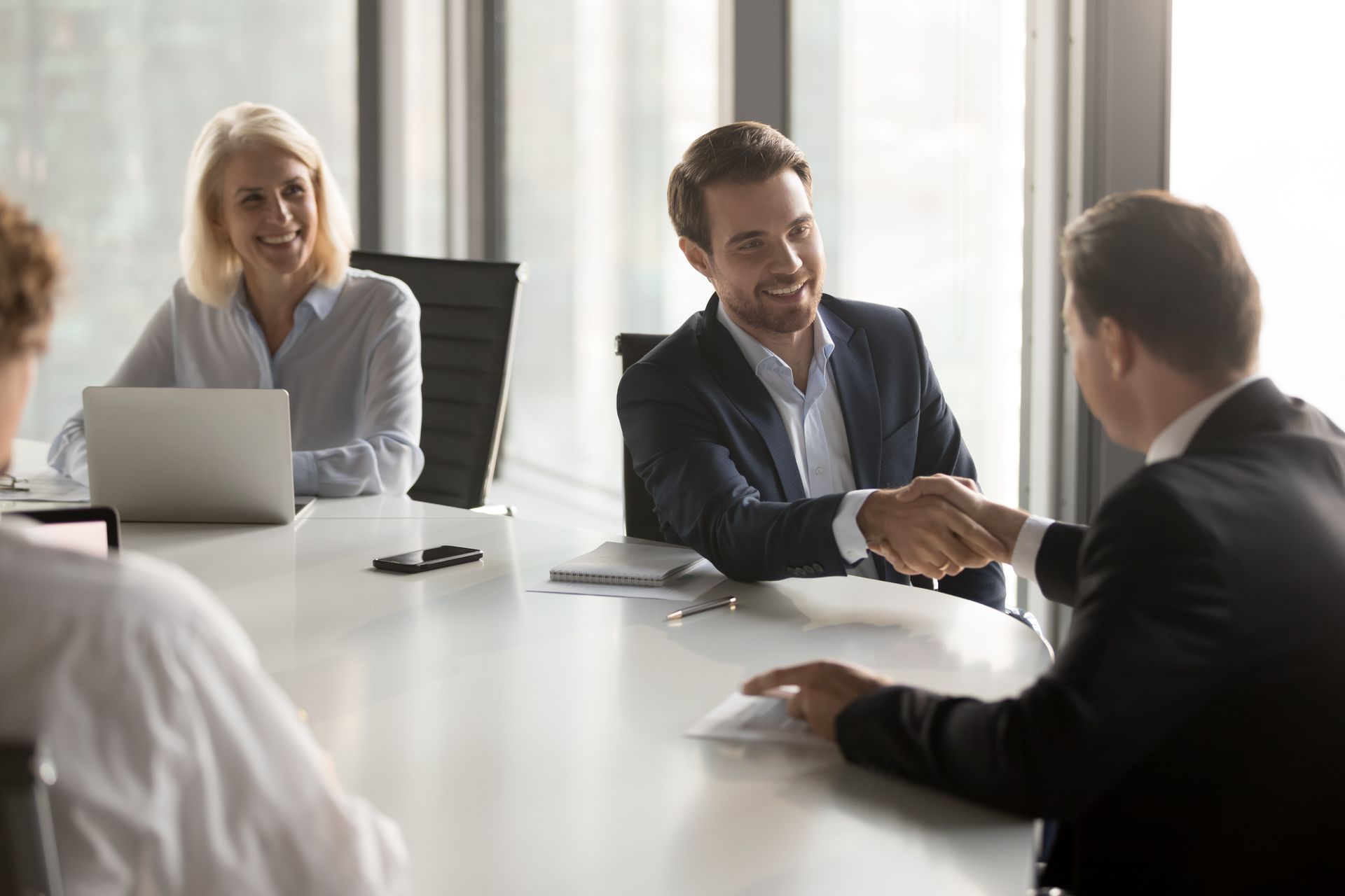 People in business attire shaking hands at a table, smiling, in a modern office.