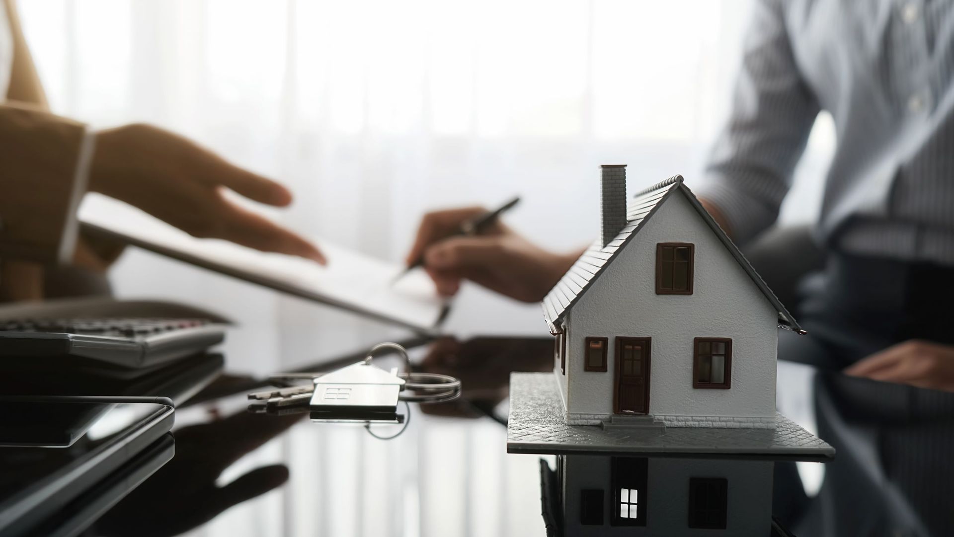 A person signing paperwork next to a miniature house model, keys, and a calculator on a reflective surface.
