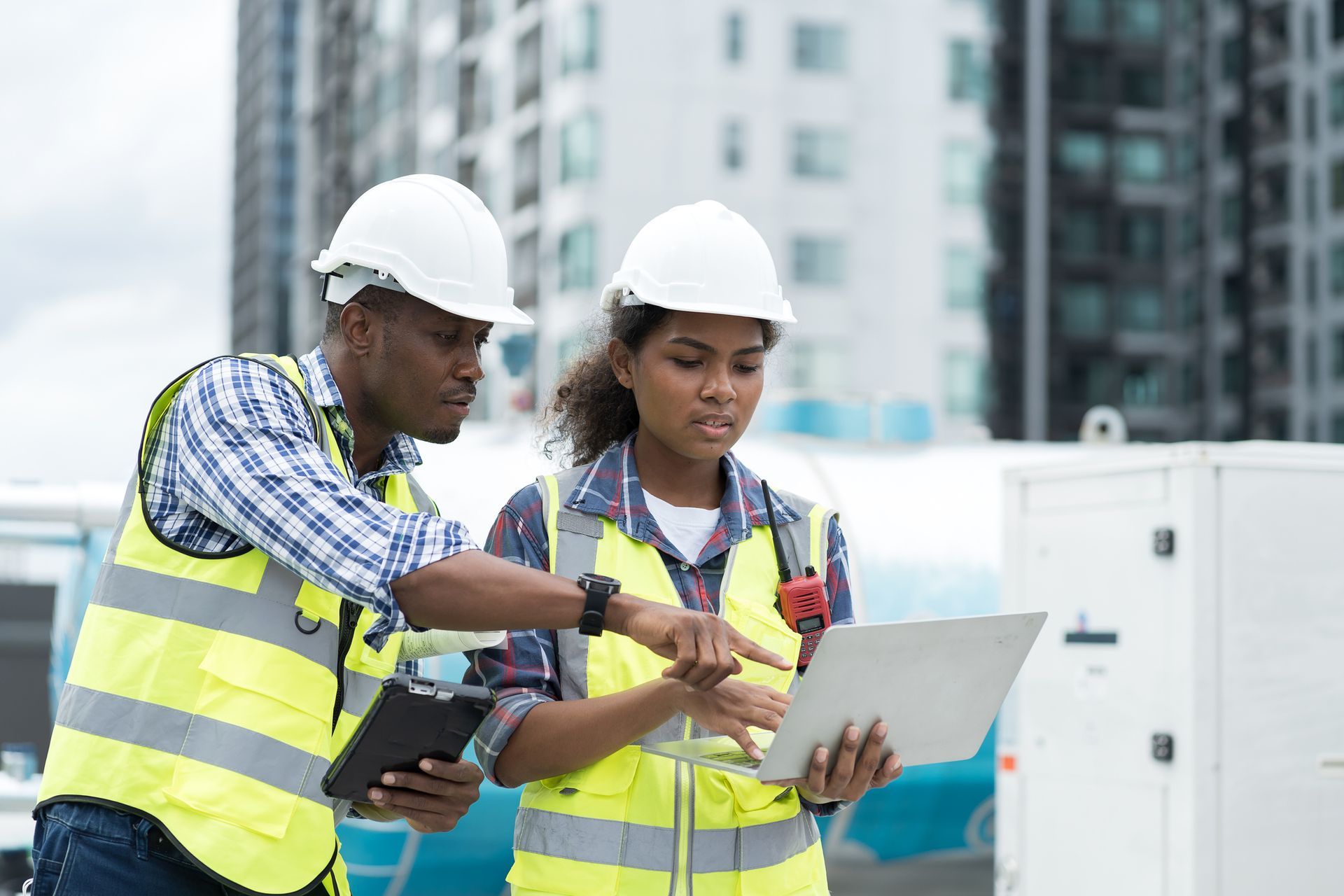 Two construction workers in safety vests and hard hats reviewing a laptop and tablet outdoors.