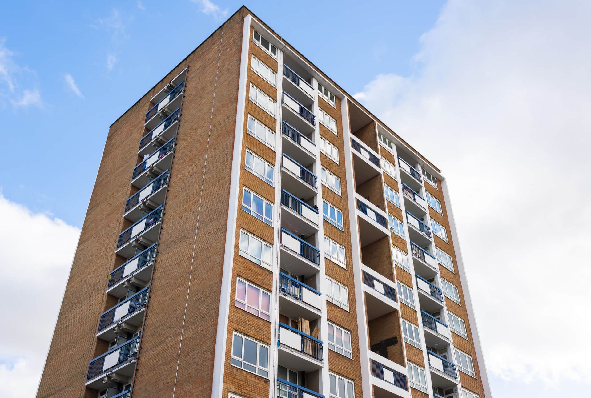 Tall brick apartment building with white window frames and blue balcony railings against a cloudy sky.