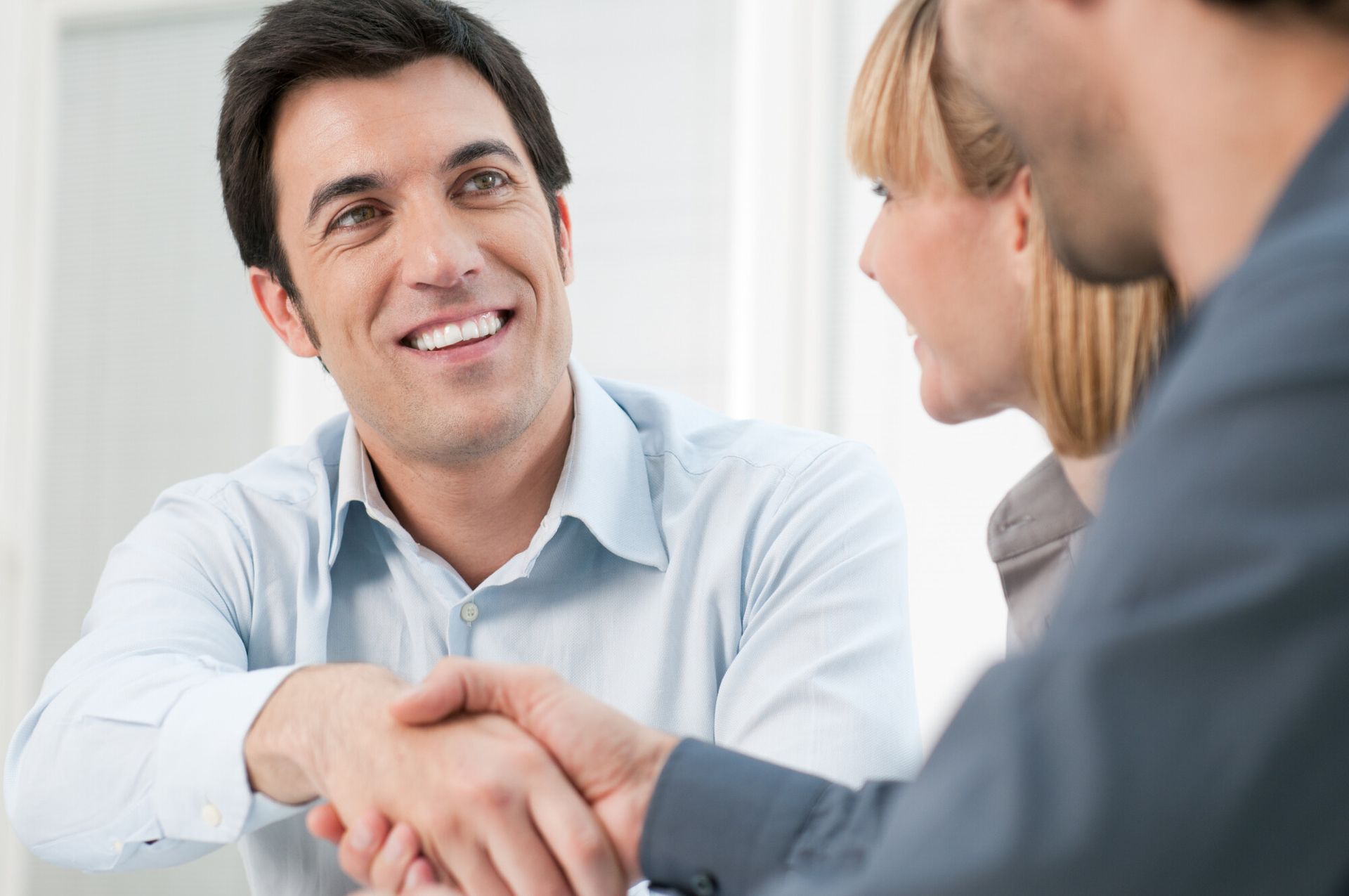 Man smiling, shaking hands with another man in an office. A woman watches.