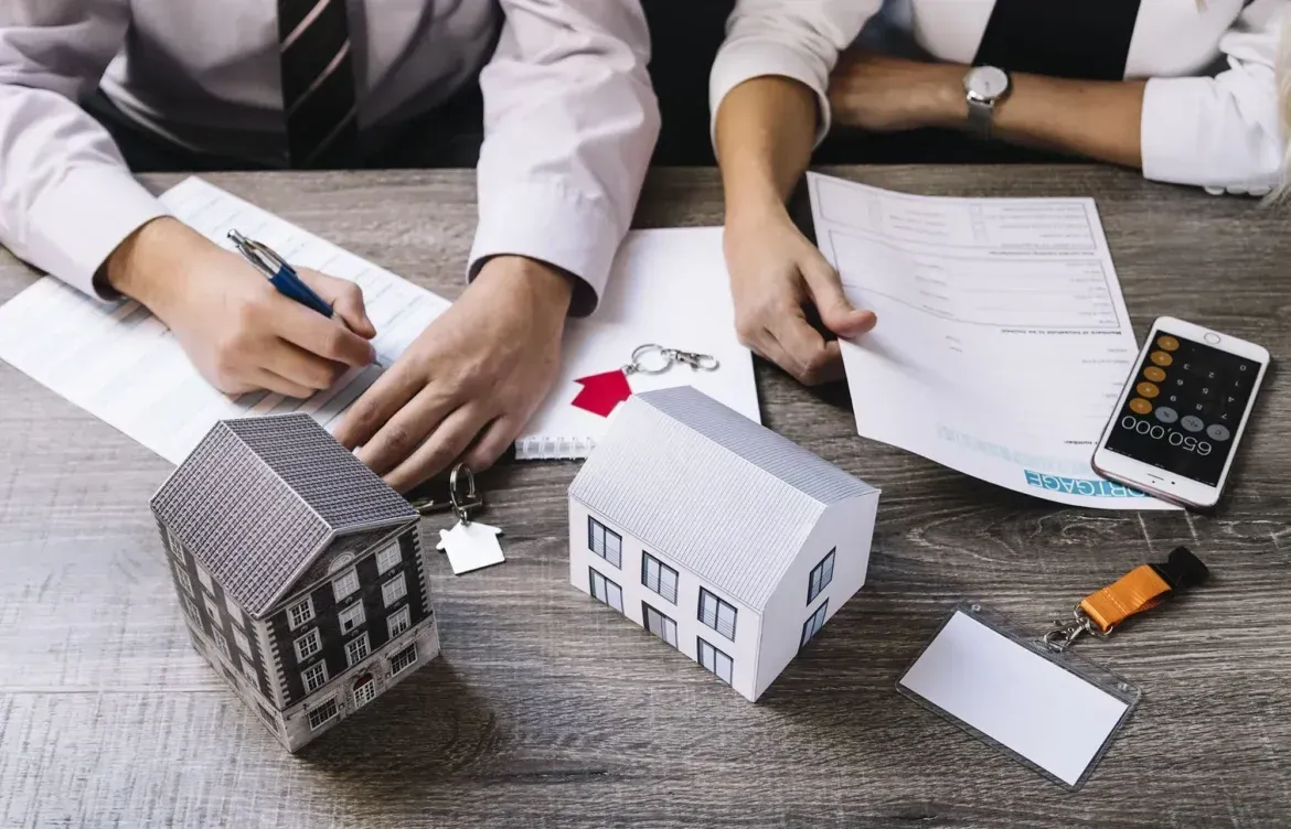 Two people at a table reviewing documents, with house models, keys, and a calculator, likely discussing a real estate deal.
