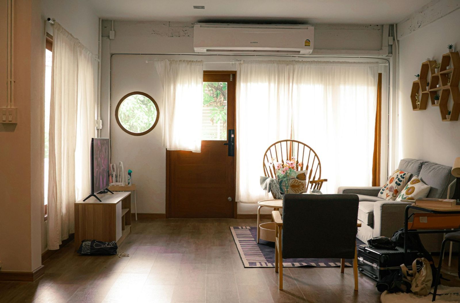 Cozy living room with wooden floors, neutral curtains, and natural light. Includes a sofa, chair, TV, and coffee table.