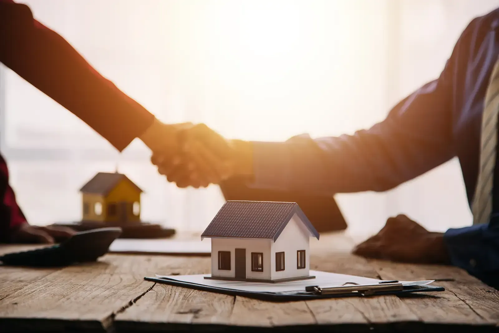 People shaking hands over a miniature house model and paperwork on a wooden table.