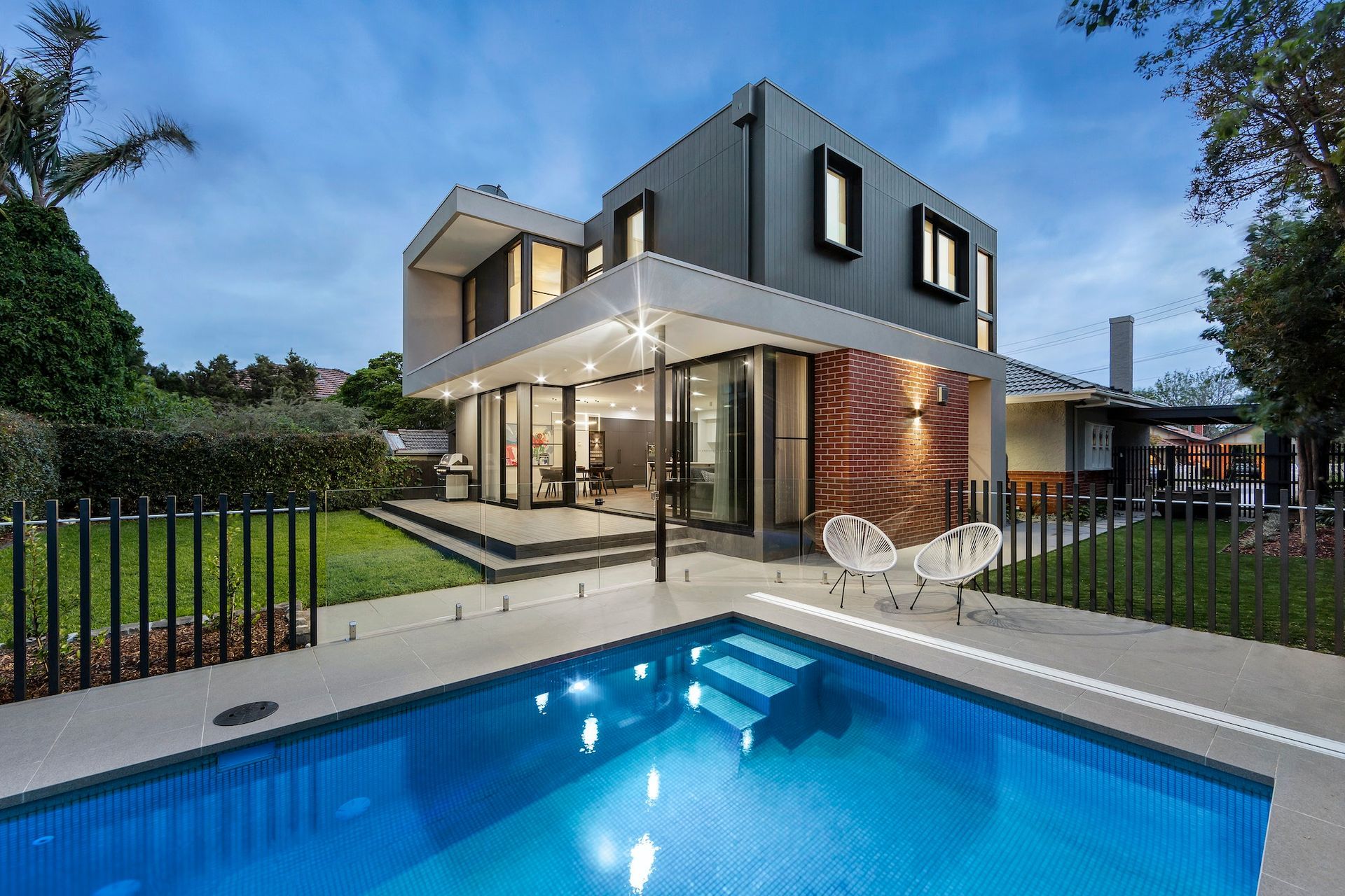 Modern home with a pool, glass doors, and a dark gray exterior, set in a grassy yard under a twilight sky.