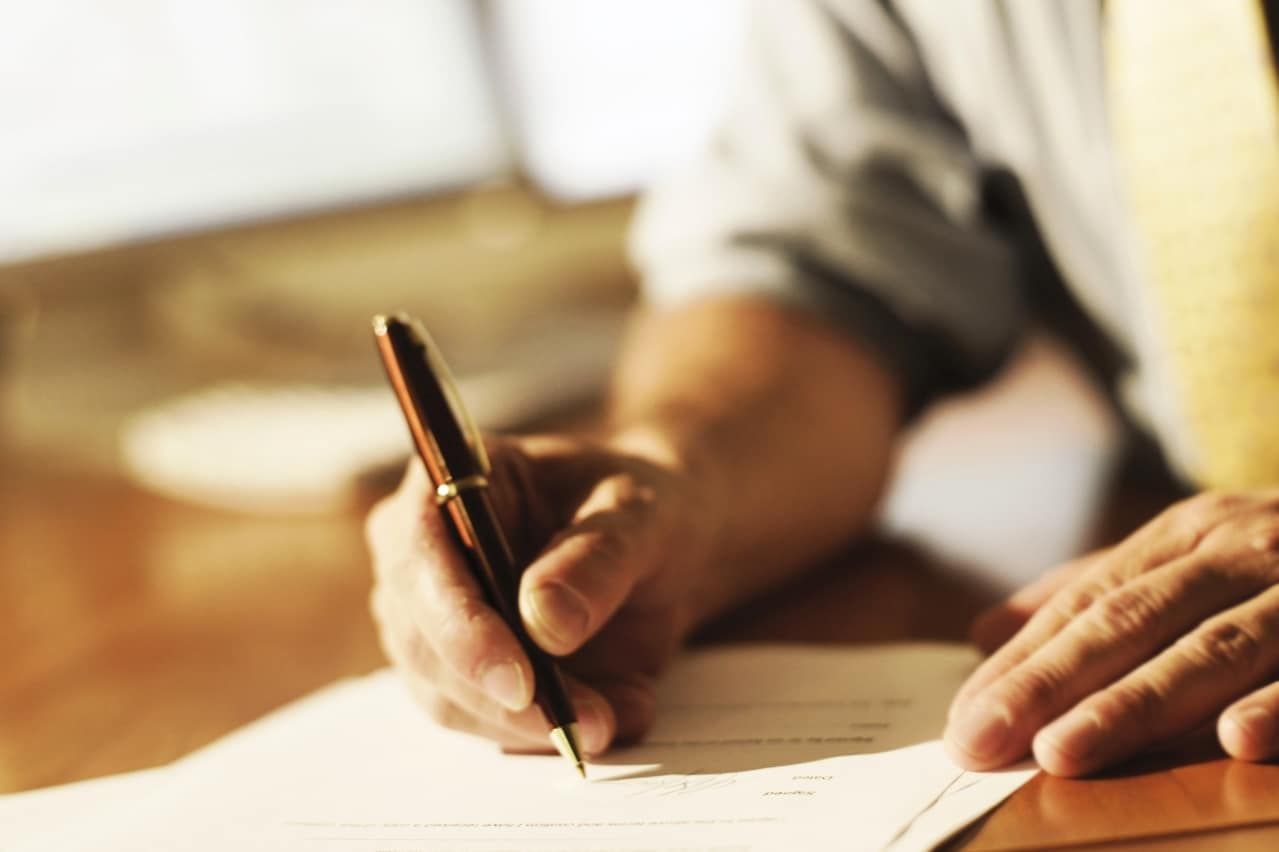 Hand holding a pen, writing on a light-colored paper, blurry background, indoor setting.