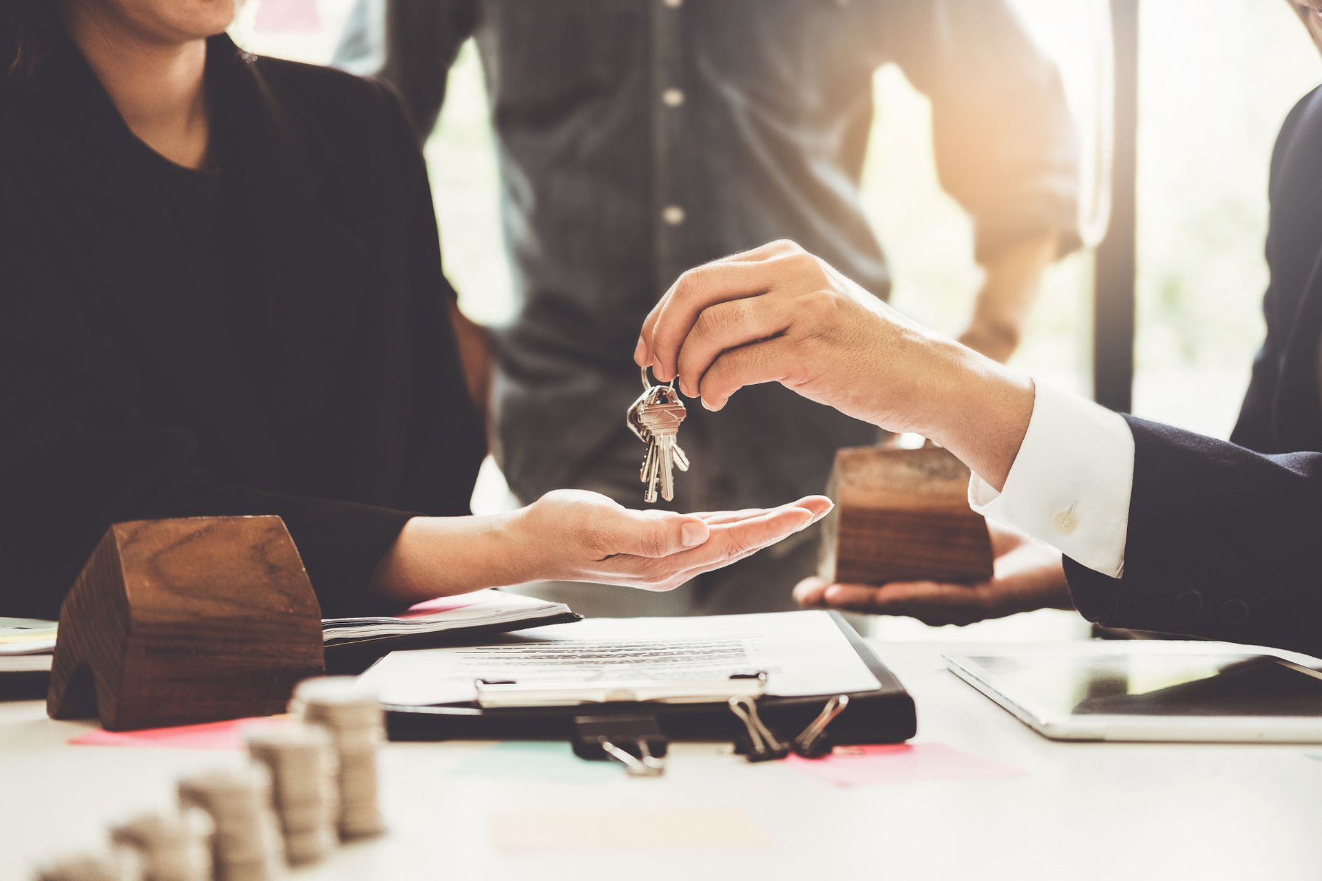 Person handing keys to another person at a table with money, miniature houses, and paperwork.