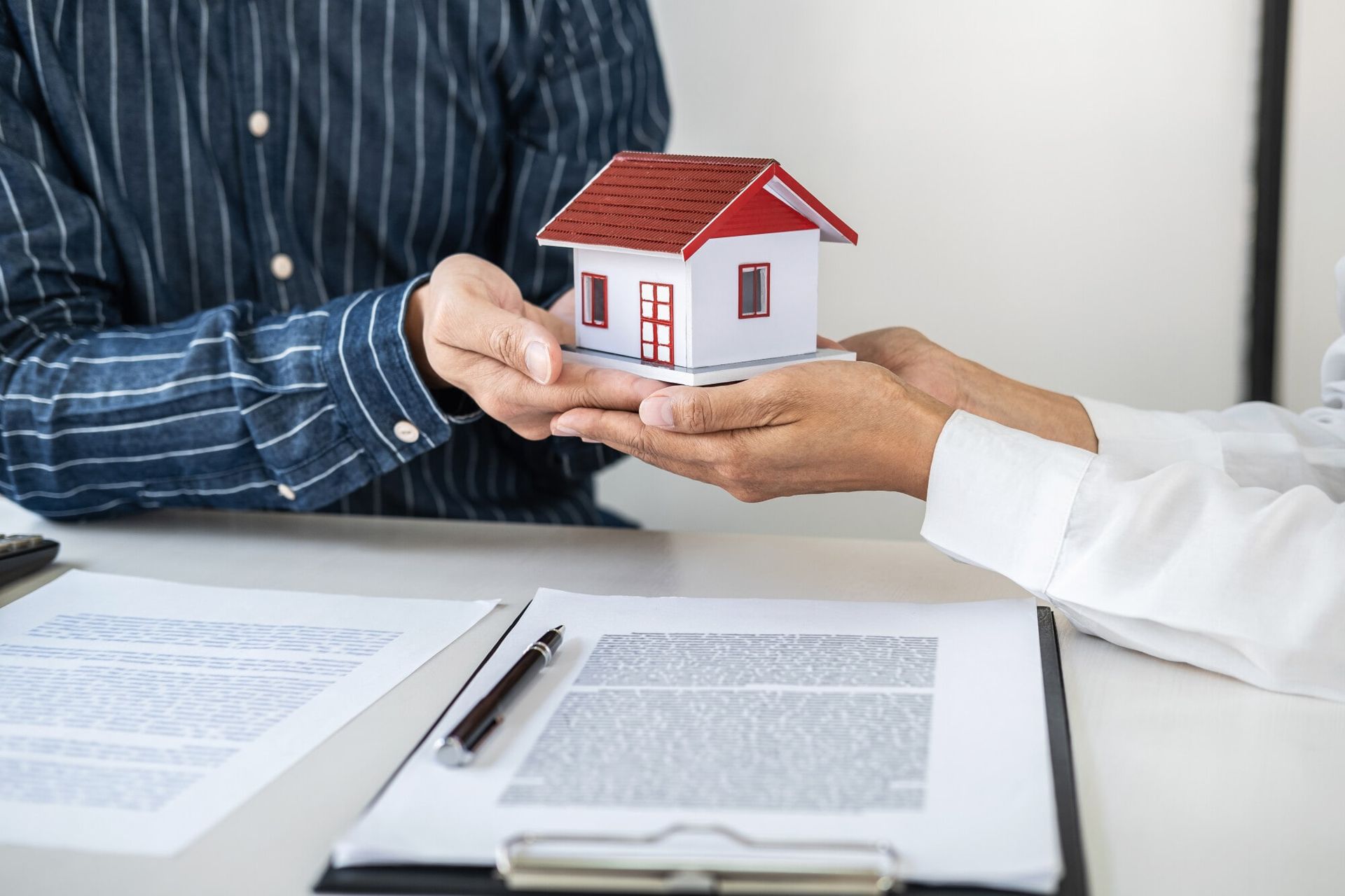 Two people exchanging a model house over documents and a pen.