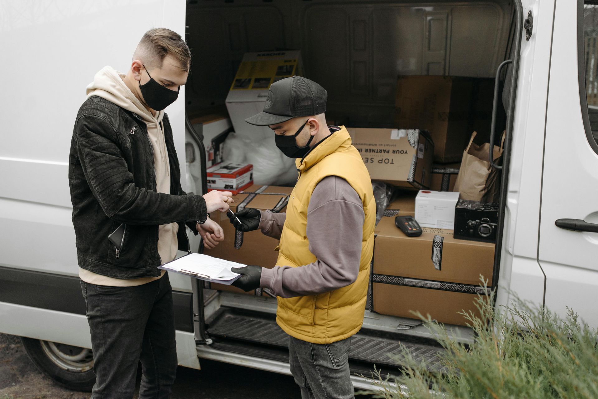 Two masked workers loading boxes into a white delivery van outdoors