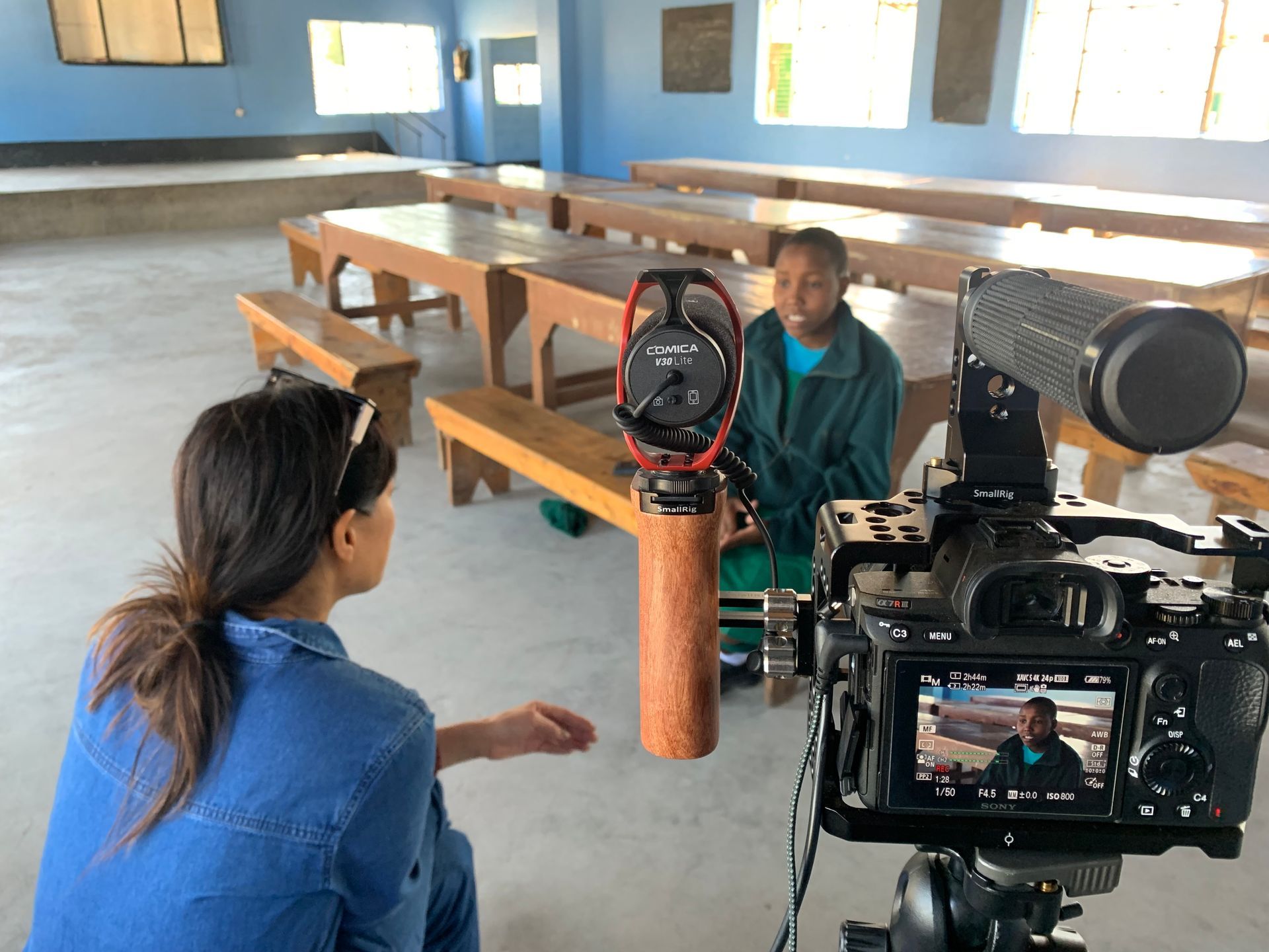 A young woman is being interviewed, another is operating a camera. They are in a room with tables and benches.