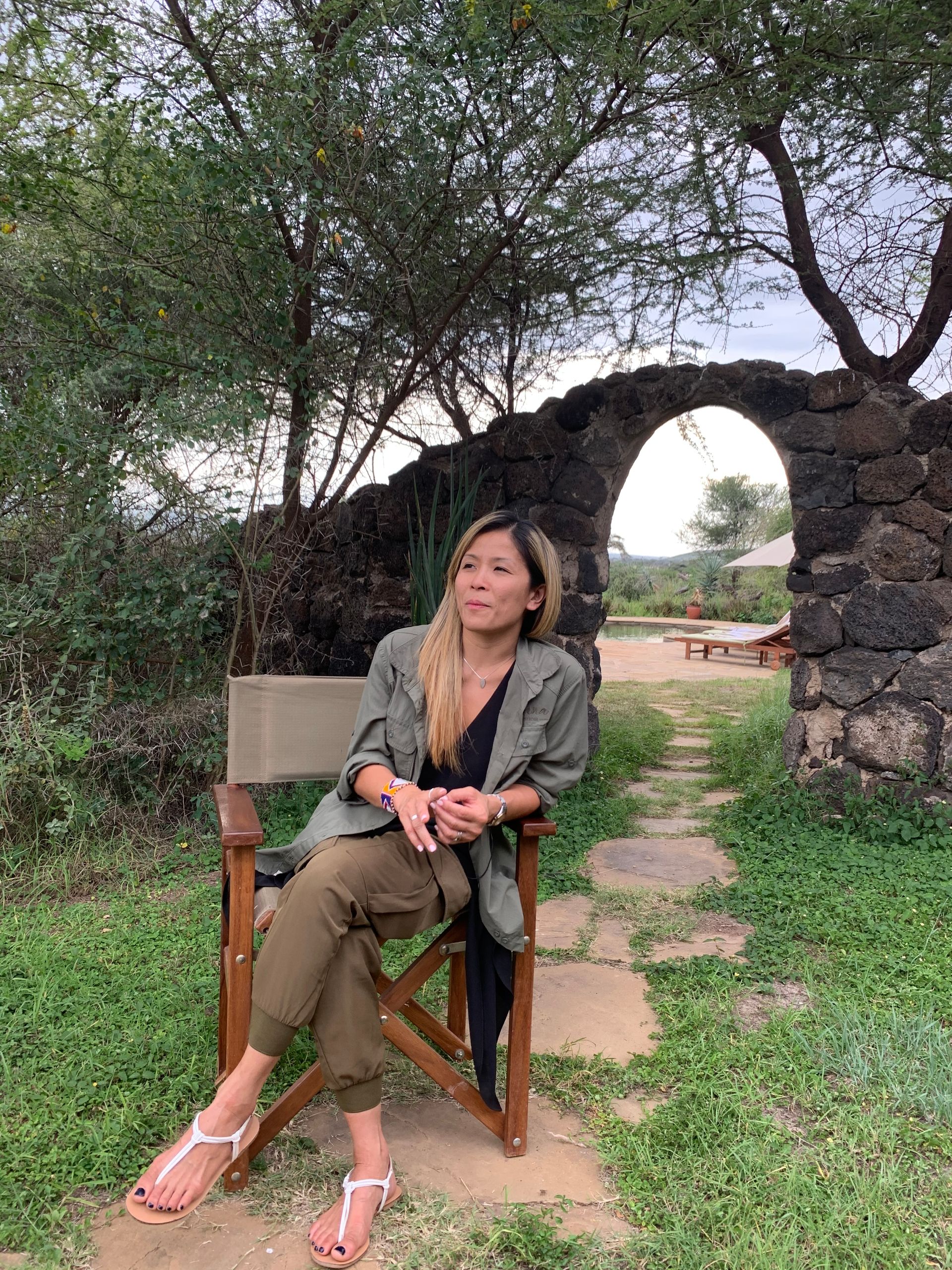 Woman seated in chair under archway, wearing olive green pants, looking off to the side.