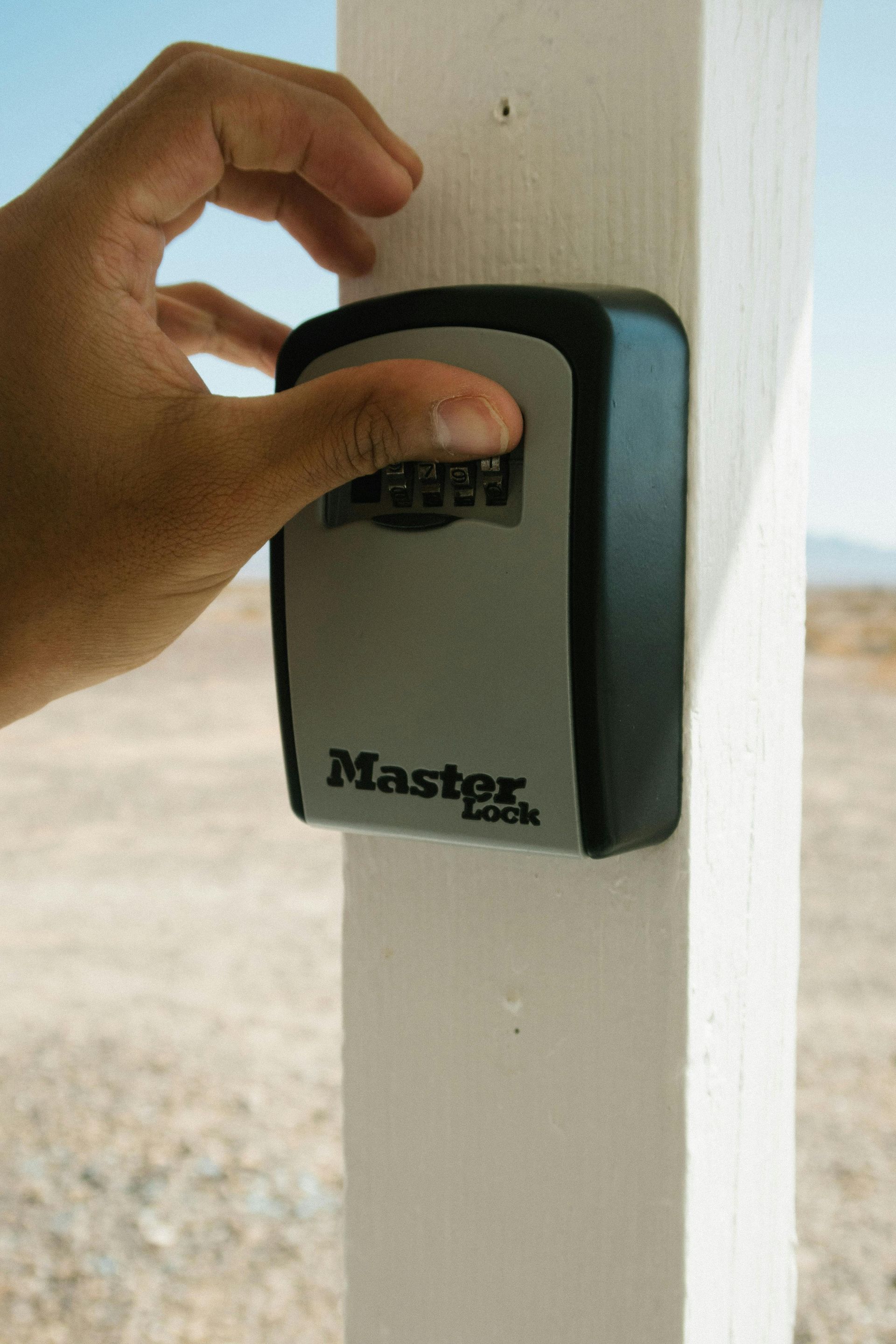 Hand pressing buttons on a Master Lock key safe attached to a white post outdoors.