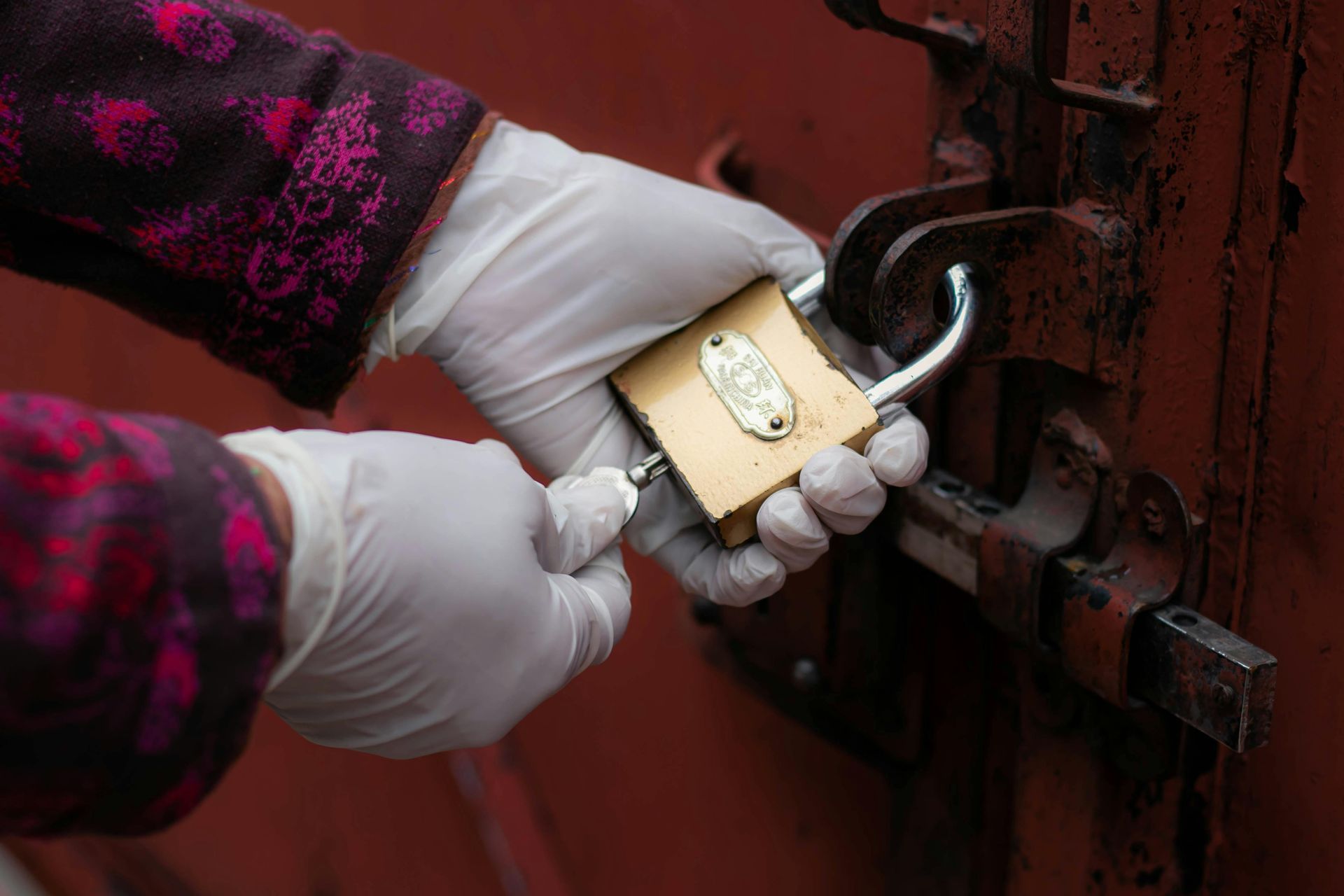 Gloved hands opening a padlock on a rusty metal door.