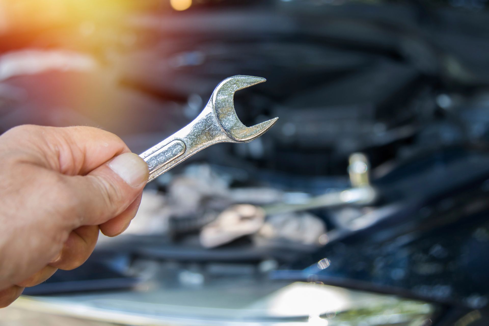 Mano che tiene una chiave inglese davanti al cofano aperto di un'auto, probabilmente intenta a riparare il motore.