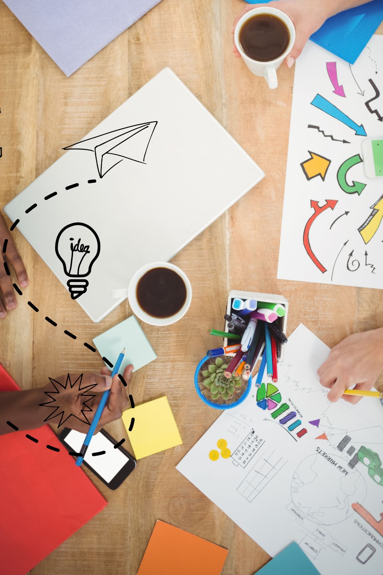 Vista aérea de una mesa de madera con una computadora portátil, bocetos, tazas de café, notas adhesivas y manos trabajando juntas.