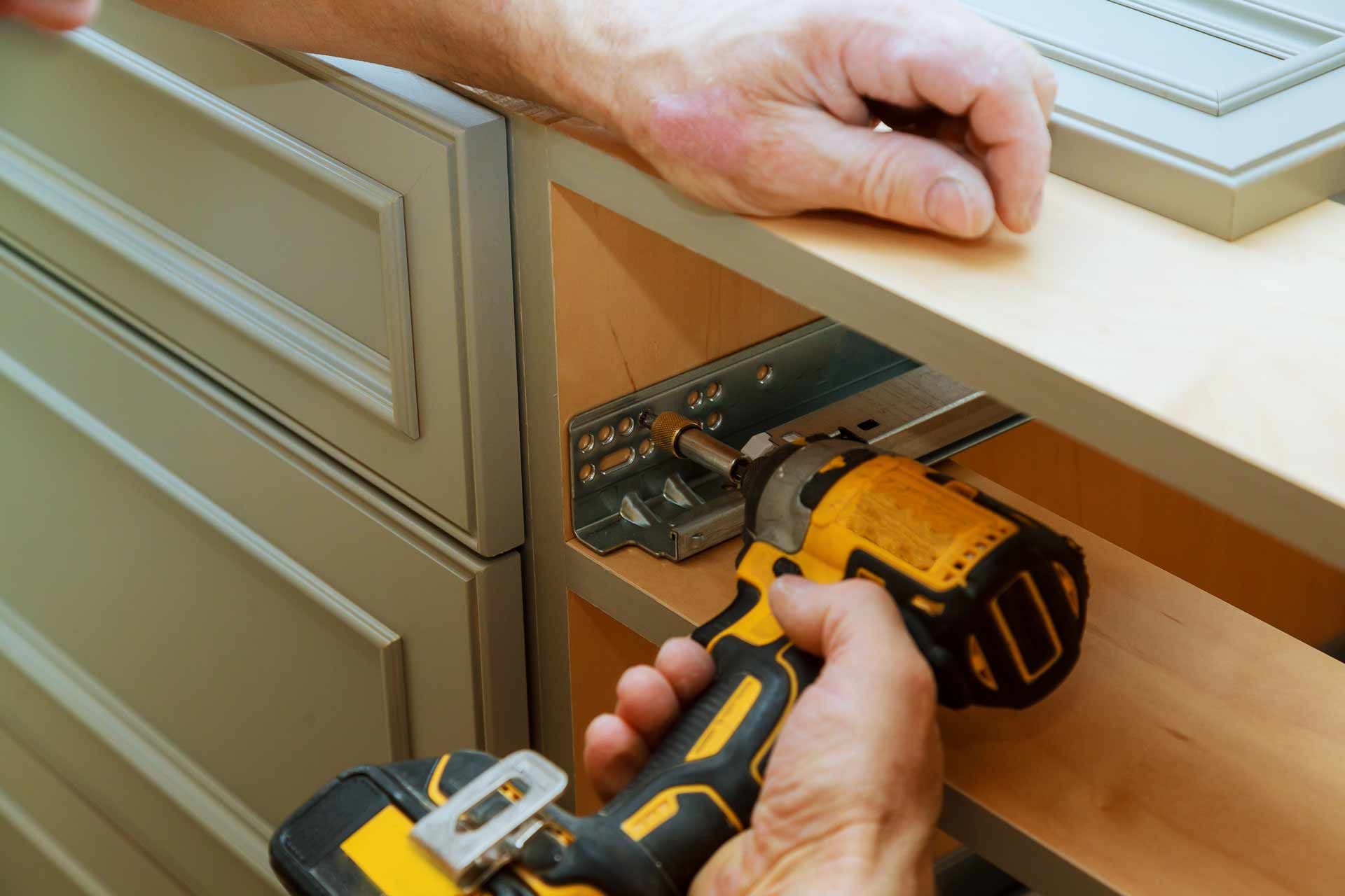 A person uses a yellow cordless drill to install a metal drawer slide into a wooden cabinet frame.