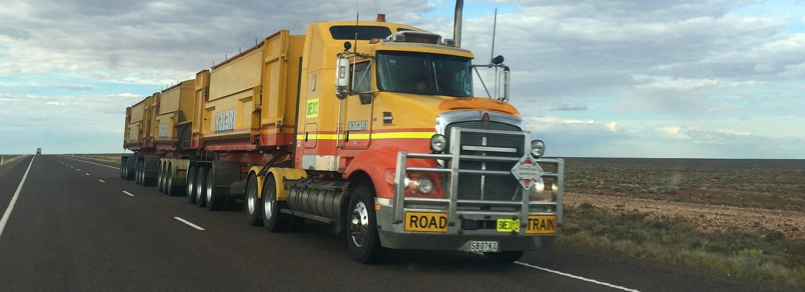 A large yellow semi-truck with multiple trailers drives on a highway, with a cloudy sky.