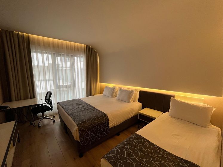 Hotel room with a king-size bed, desk, TV, and large window with curtains. Modern design with gray and orange accents.