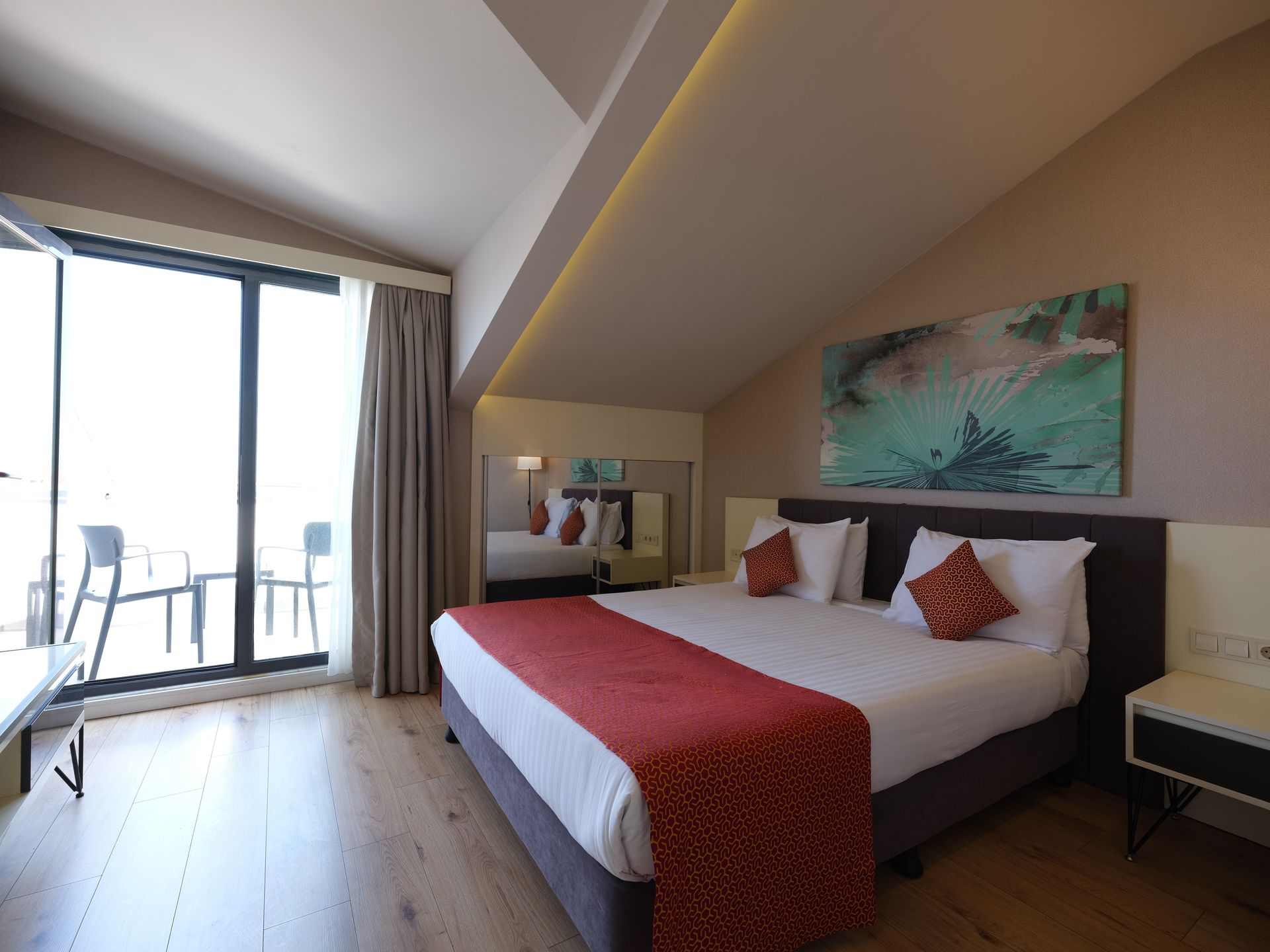 A hotel bedroom with a bed, balcony, and artwork; brown, red, and white decor. Angled ceiling and wooden floor.