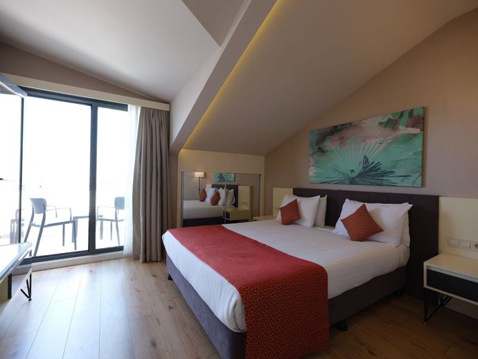 A hotel bedroom with a bed, balcony, and artwork; brown, red, and white decor. Angled ceiling and wooden floor.