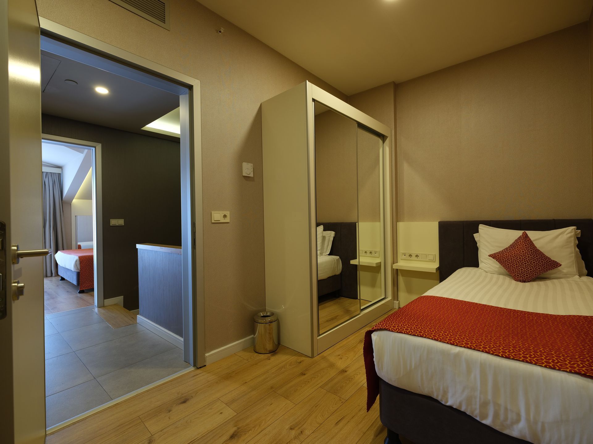 A hotel room with a single bed, a wardrobe, and an open doorway leading to another room with a bed.