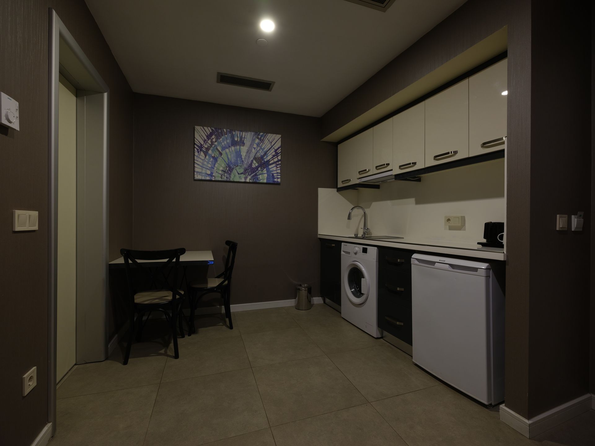 Small kitchen area with appliances, cabinets, a table, and chairs against a dark wall. A painting hangs above the table.