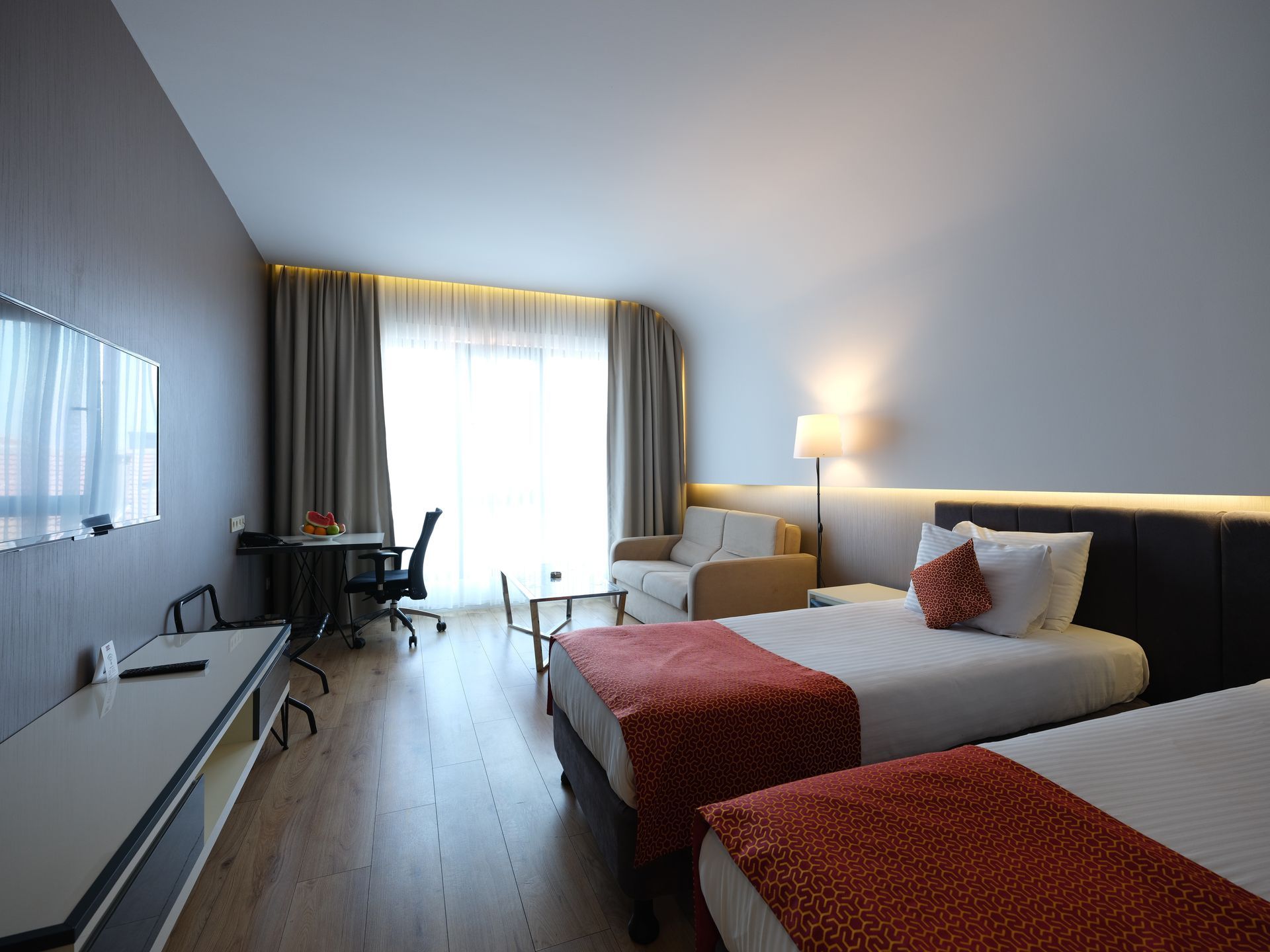 A hotel room with two twin beds, a couch, desk, and a large window with drapes; warm lighting and wooden floors.