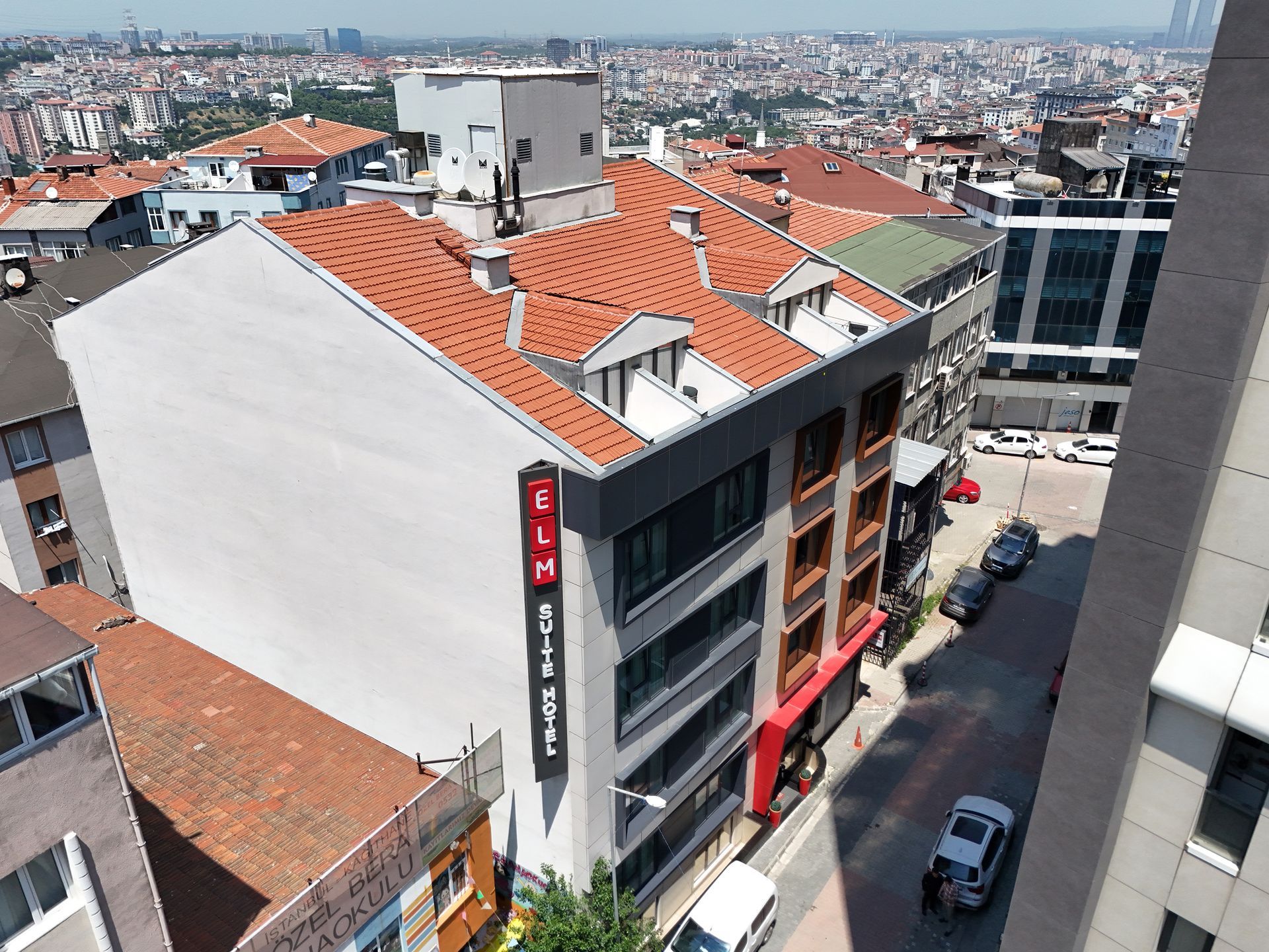 Modern building with red tile roof and ELM HOTEL sign on a street in a city, with a cityscape in the background.