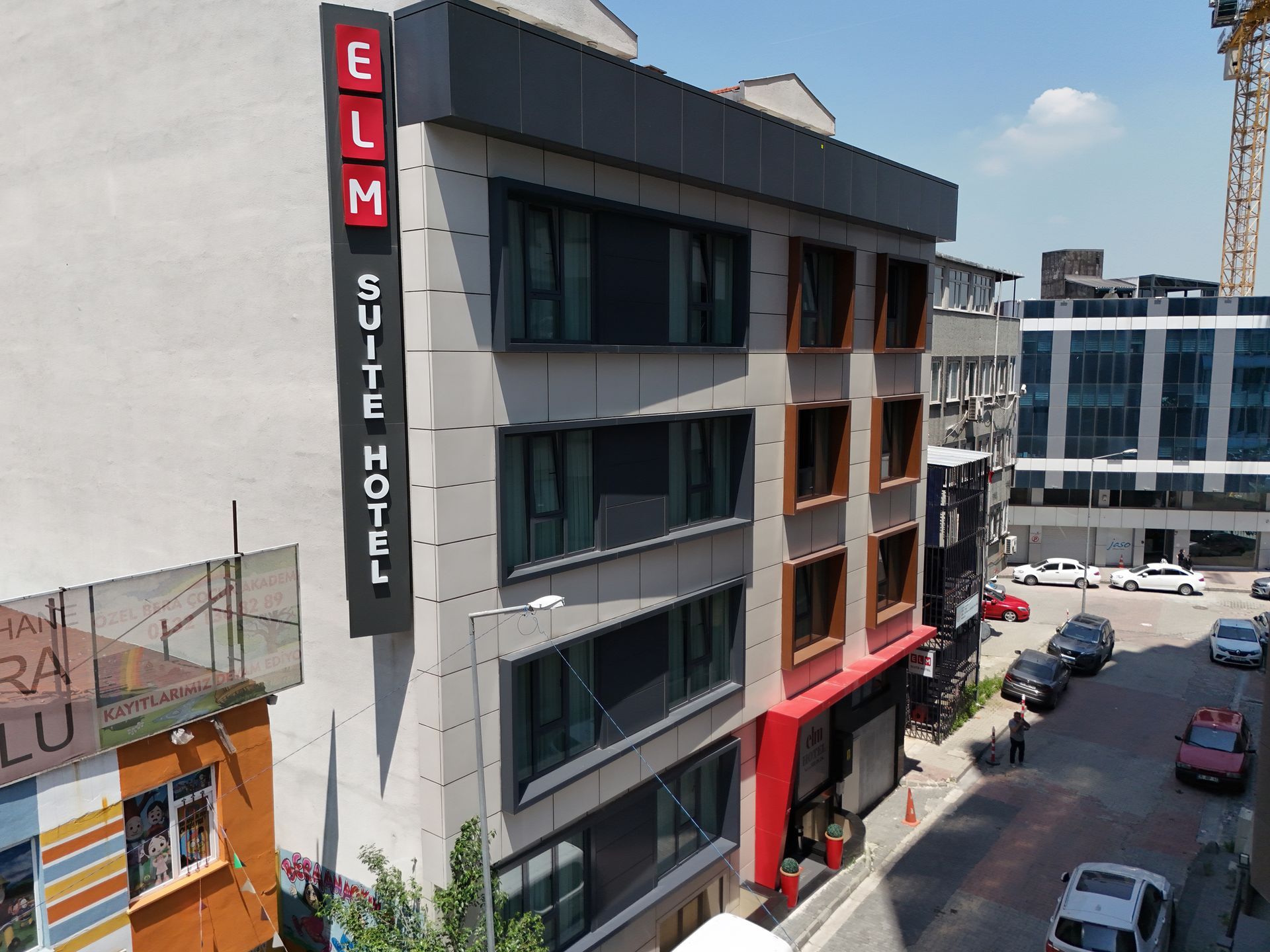 Elm Suite Hotel, a modern building with a red and gray facade, has a vertical sign. A street with parked cars is in front.