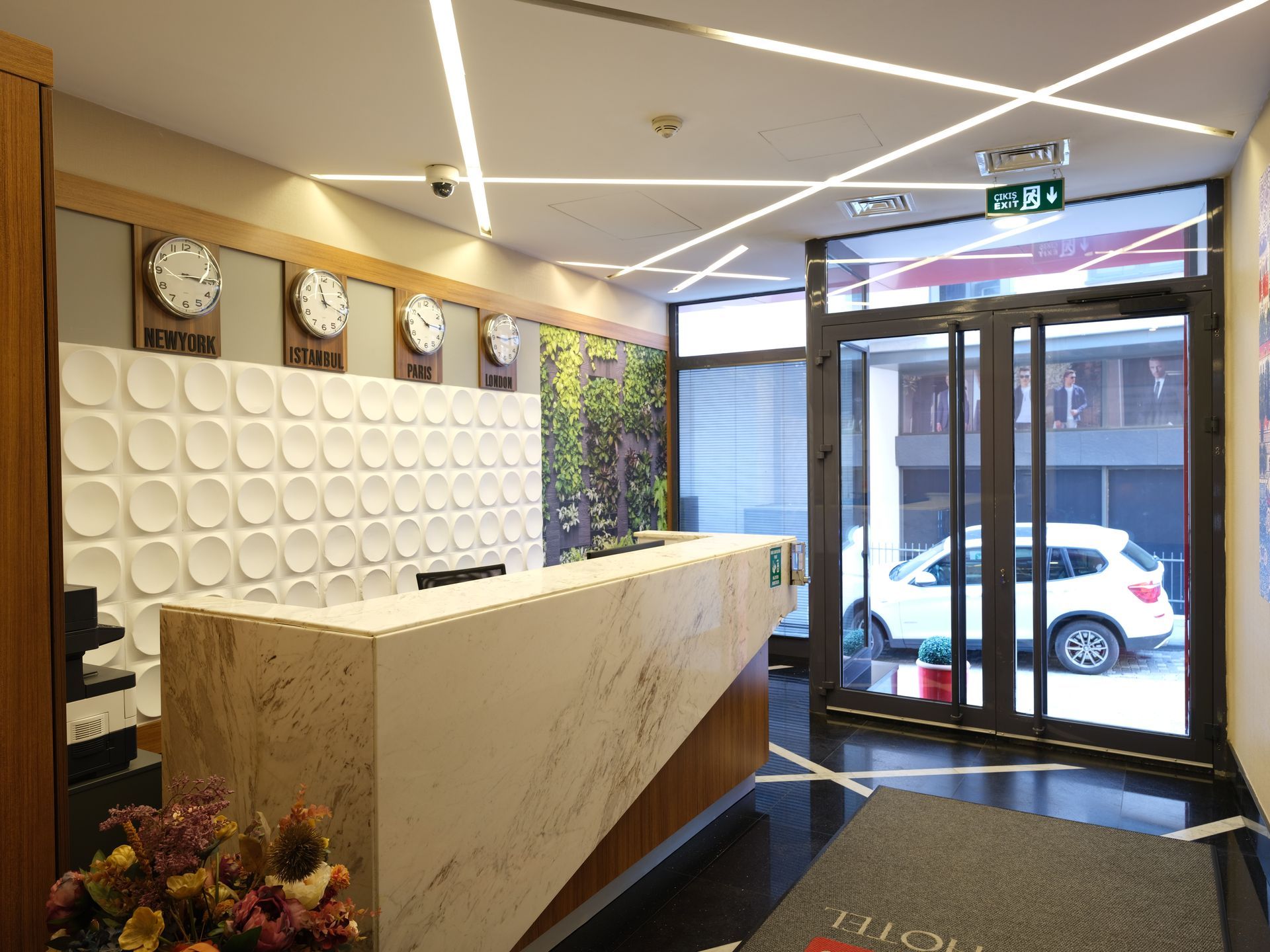 Hotel lobby with a marble reception desk, world clocks on the wall, and a glass door leading outside with a car visible.