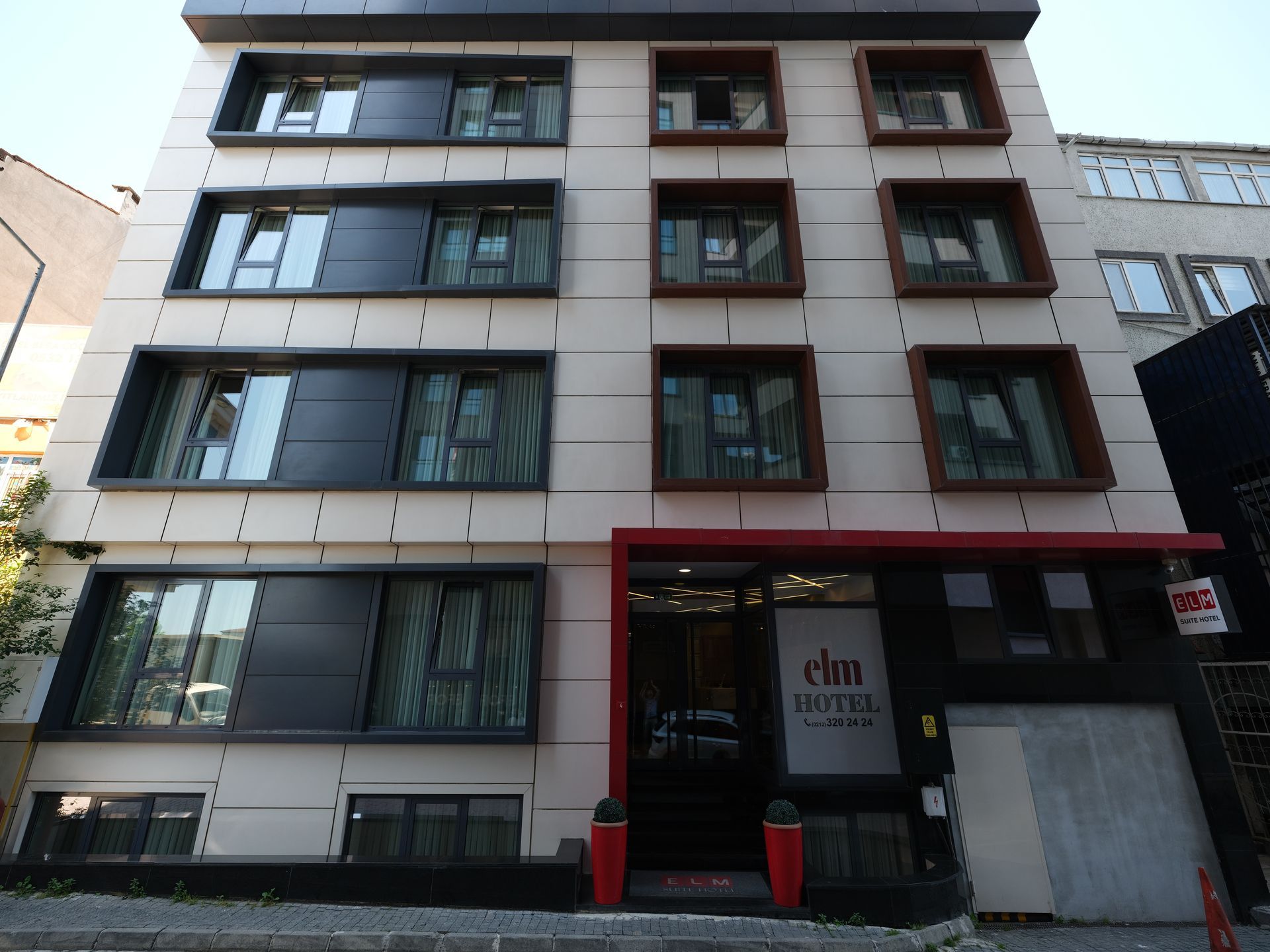 A modern hotel building with a beige facade, dark window frames, and a red entrance.