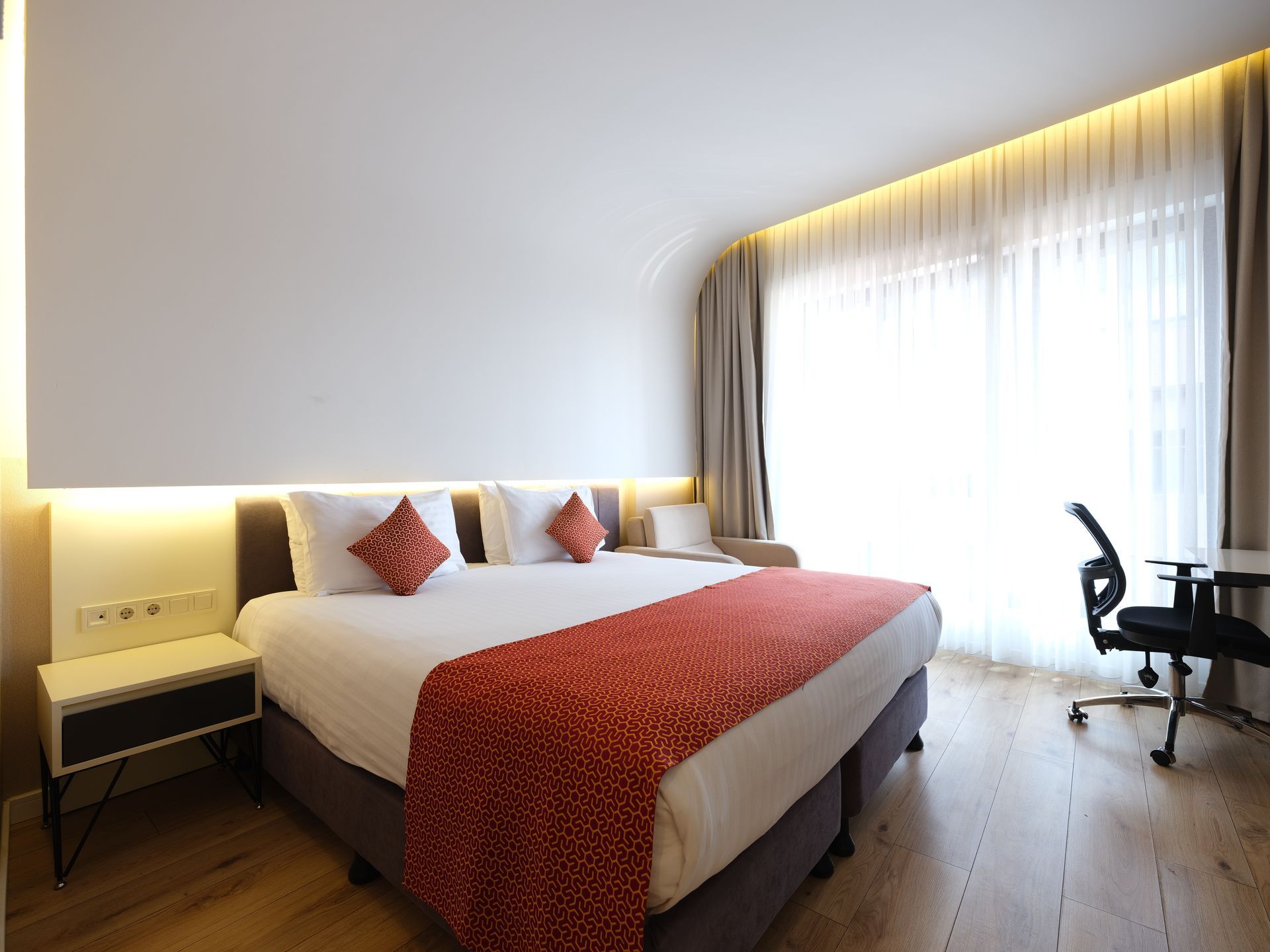 A modern hotel room with a bed, small sofa, desk, and window with sheer curtains. 