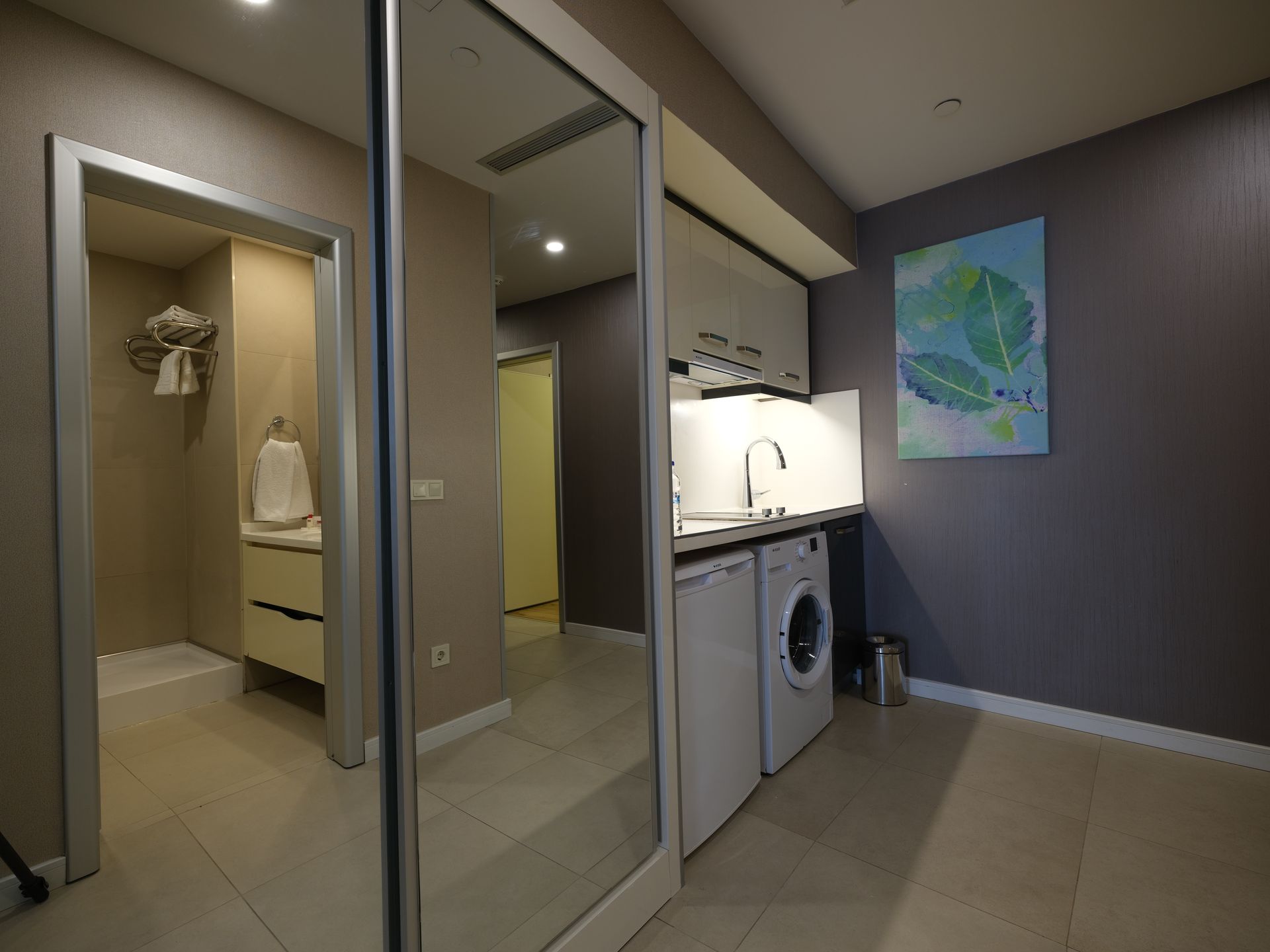 A small, modern laundry room with a washer, sink, and artwork, reflecting in a large mirror. 