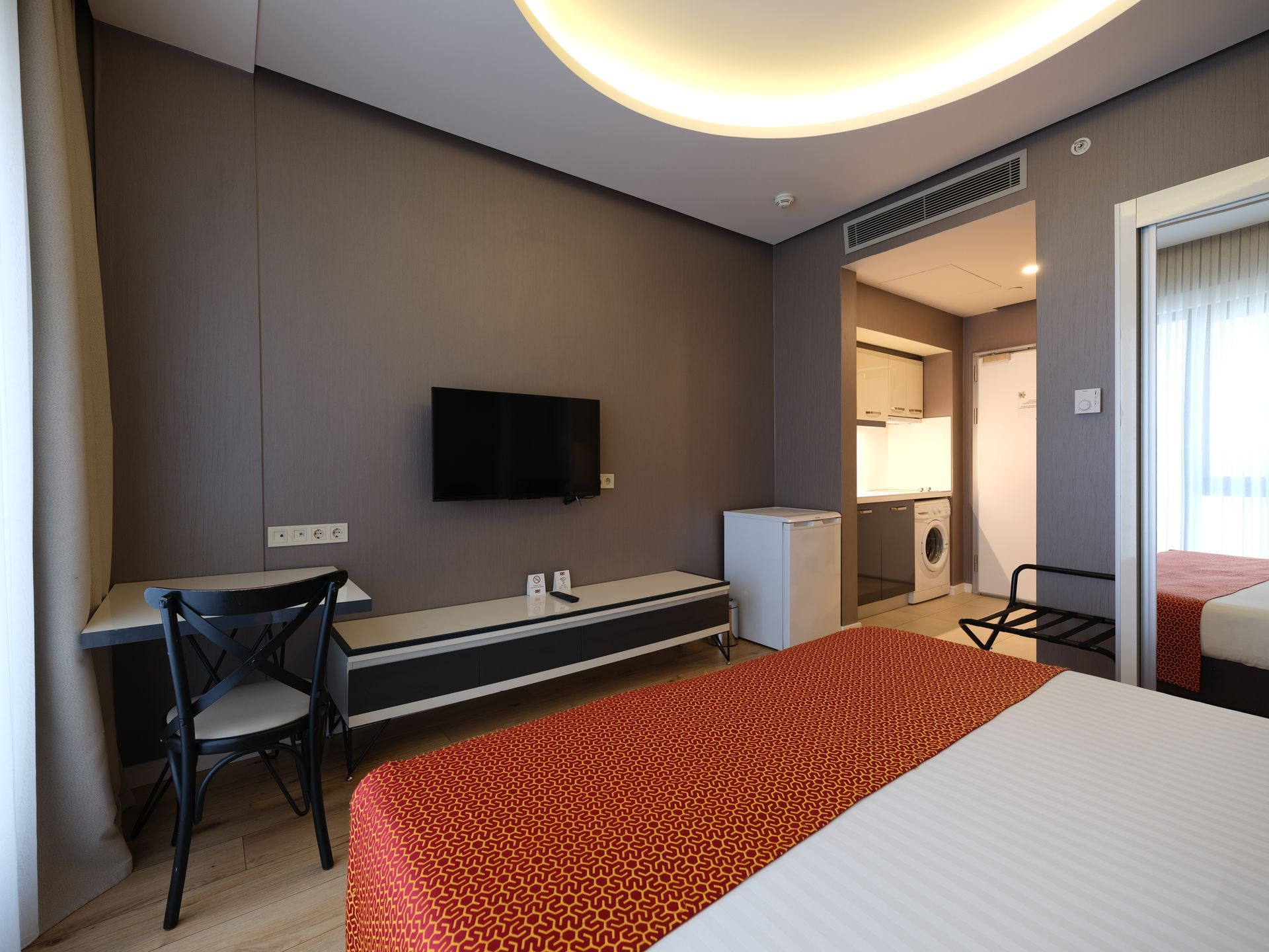 Hotel room with bed, desk, TV, and kitchenette in the background. The walls are gray with a white ceiling.