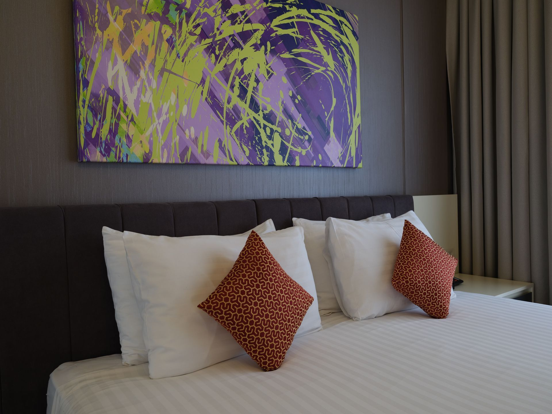 A bed with white pillows and patterned throw pillows, under a colorful abstract painting.