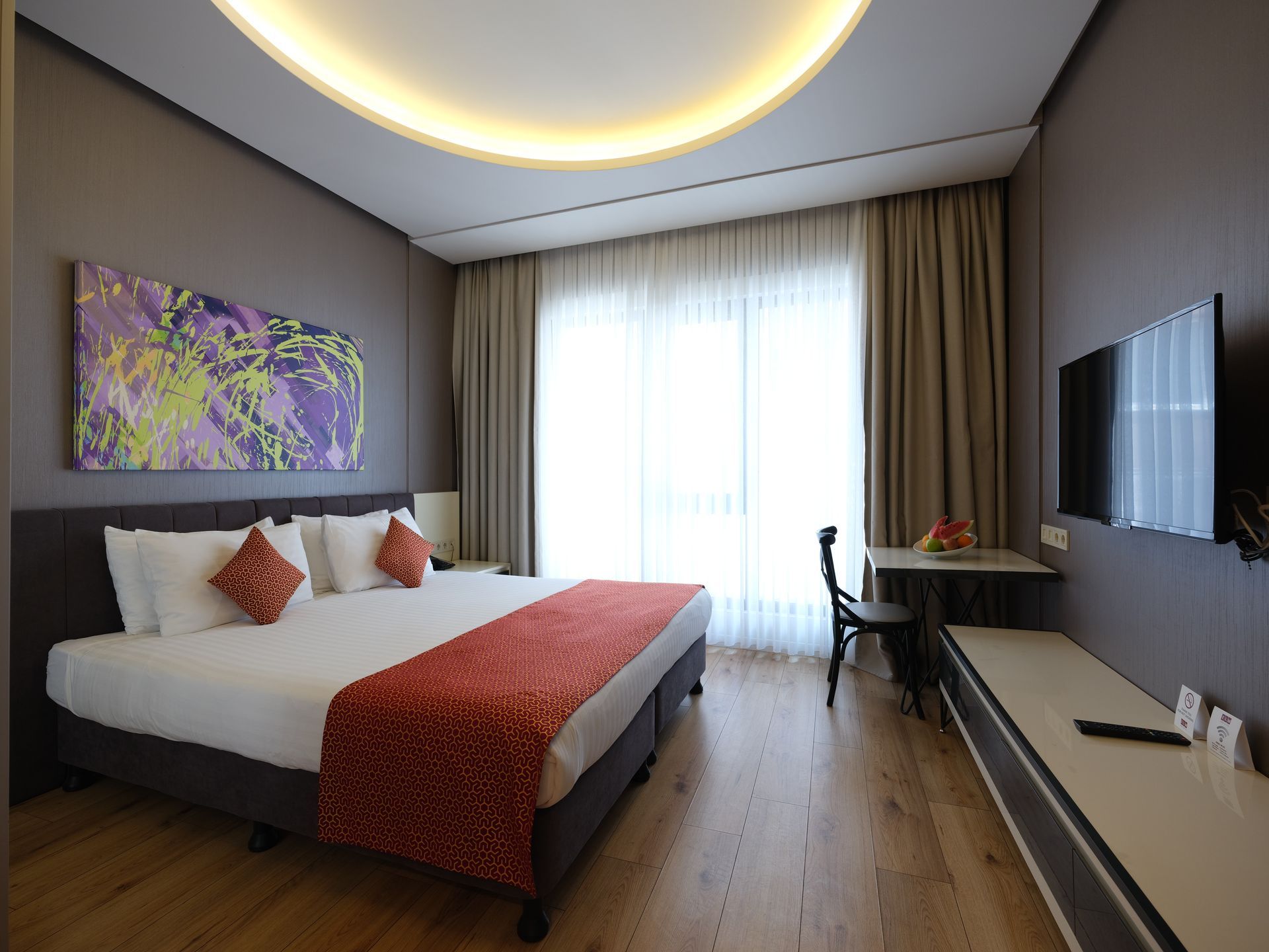Hotel room with a king-size bed, desk, TV, and large window with curtains. Modern design with gray and orange accents.