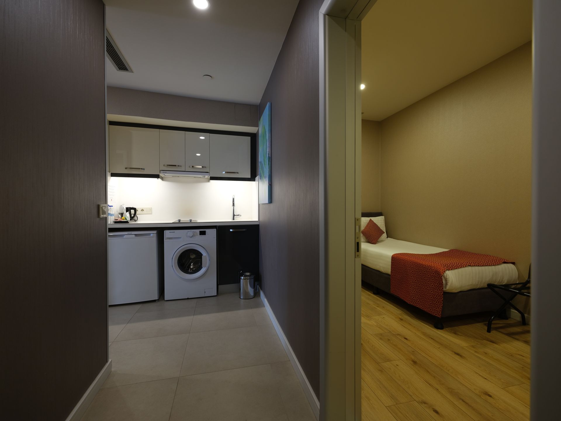 Narrow hallway leading to a kitchen on the left and a bedroom on the right. The kitchen has white cabinets and appliances.