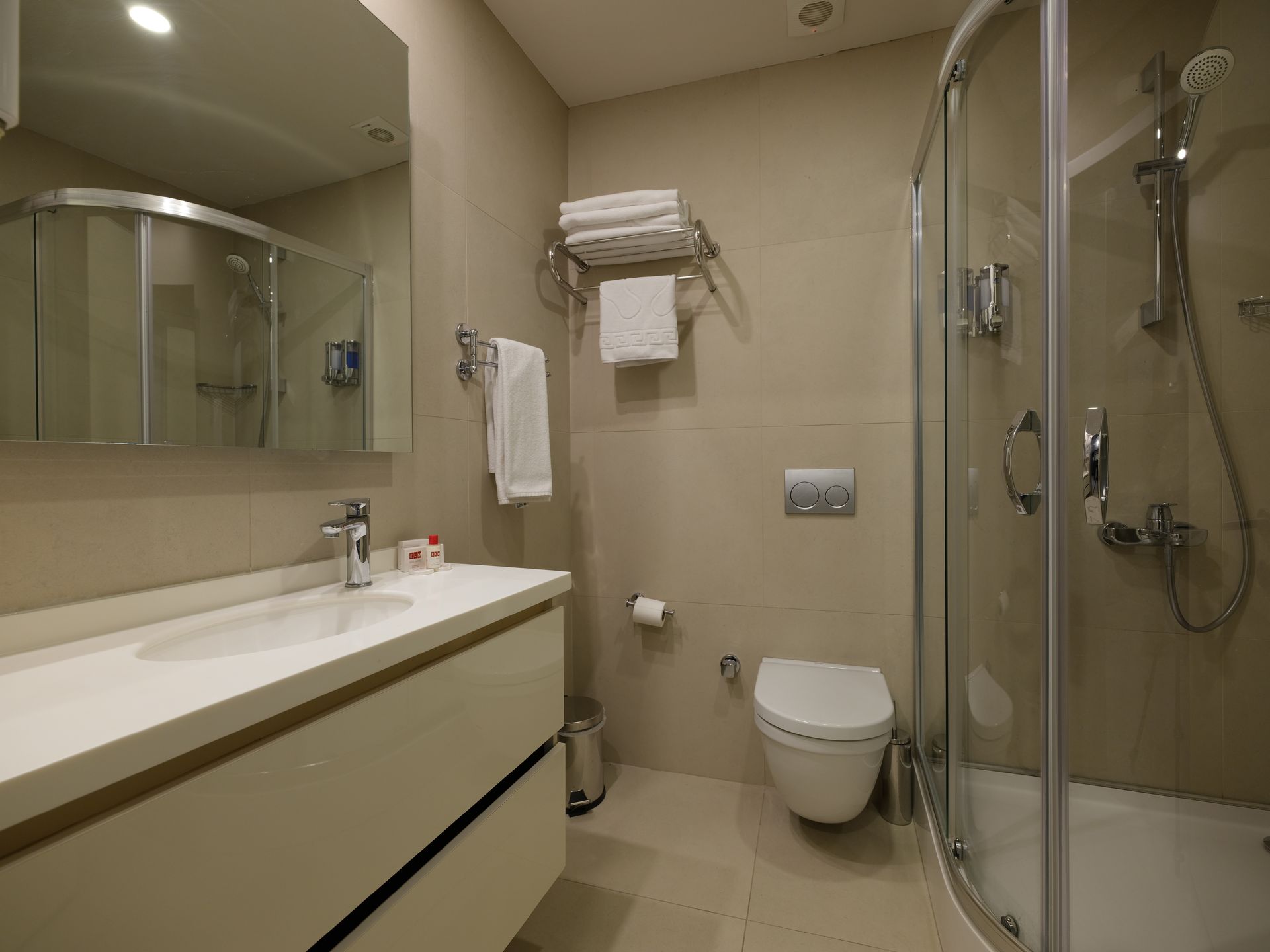 Modern bathroom with a white vanity, toilet, and a glass shower. Towels hang on a rack above the toilet.