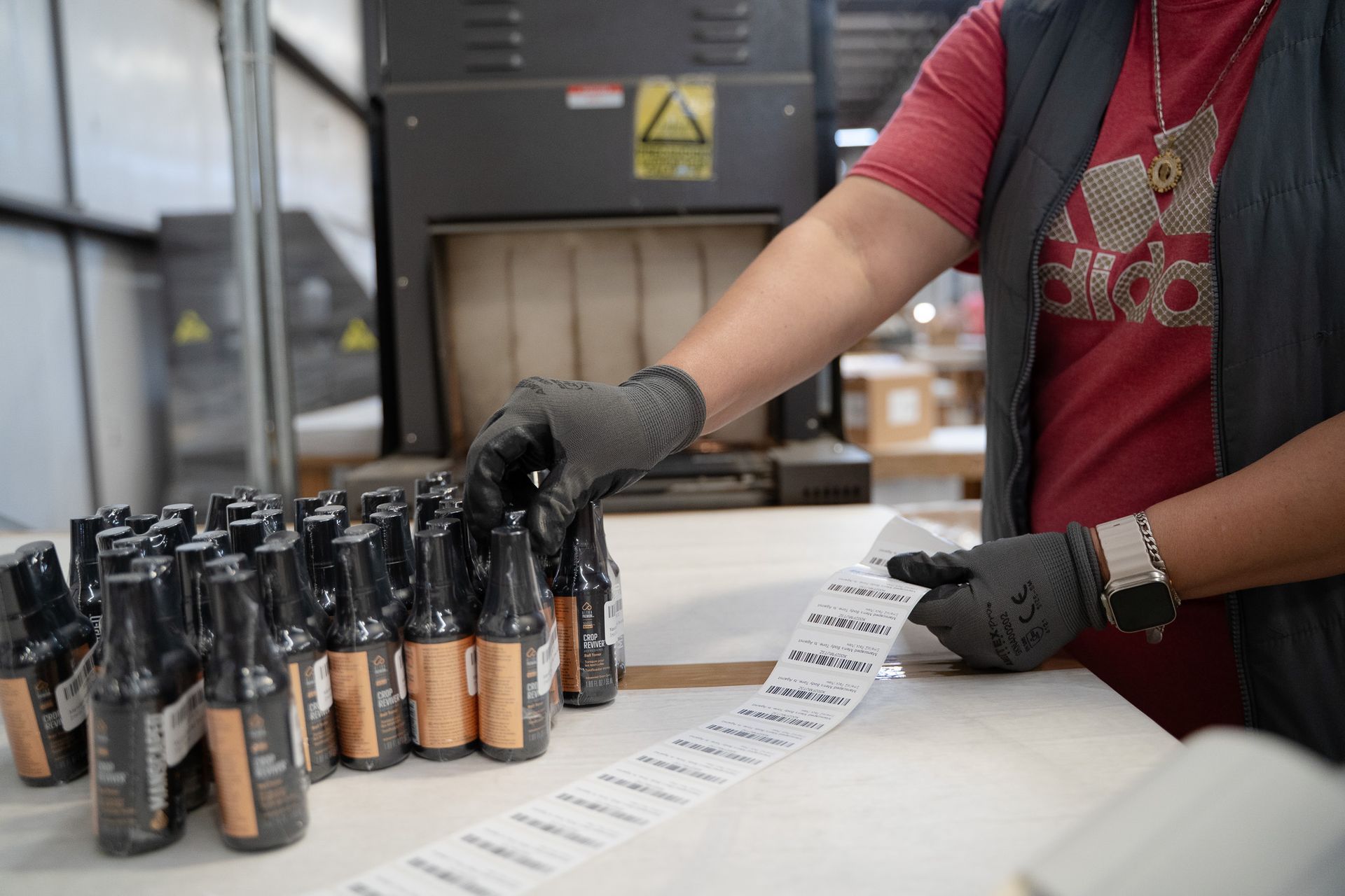 Gloved worker labels cosmetic bottles beside a printer in a factory setting.