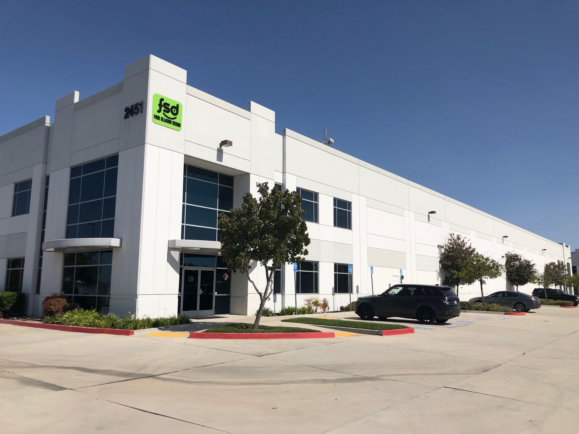 Modern white office building with a green sign, trees, and parked cars on a sunny day