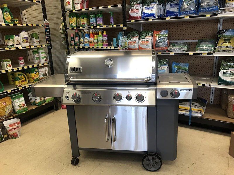 Stainless steel gas grill on display in a store, surrounded by shelves of gardening supplies.