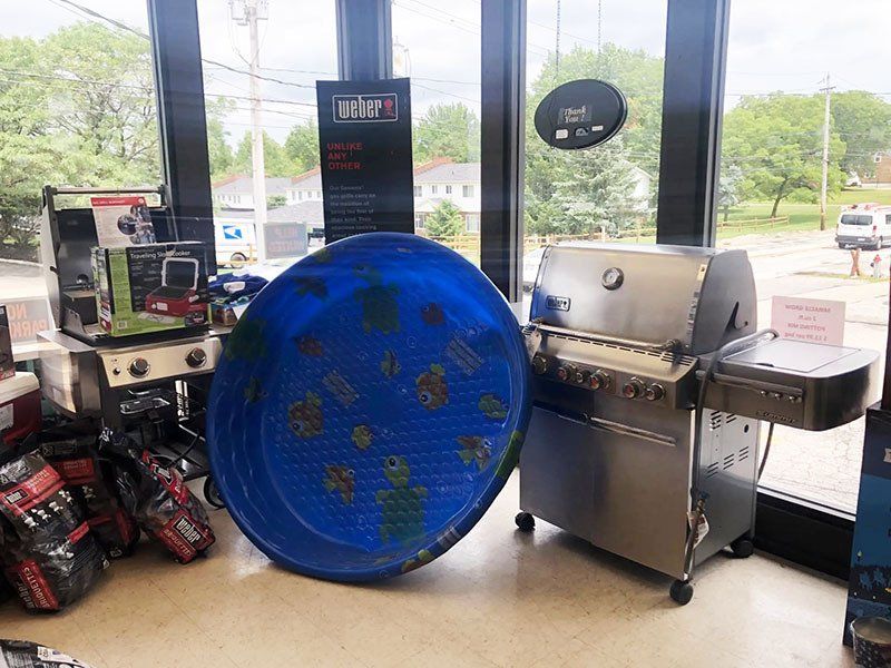 A store window displays a blue grilling mat, stainless steel grill, and charcoal bags.