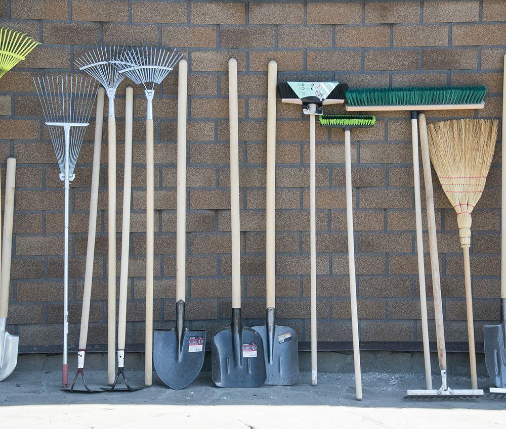 Gardening tools, including rakes, shovels, and brooms, leaning against a brick wall.