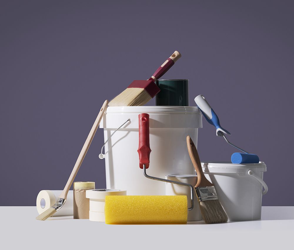 Paint supplies arranged on a white surface, including buckets, brushes, rollers, and tape, against a purple background.