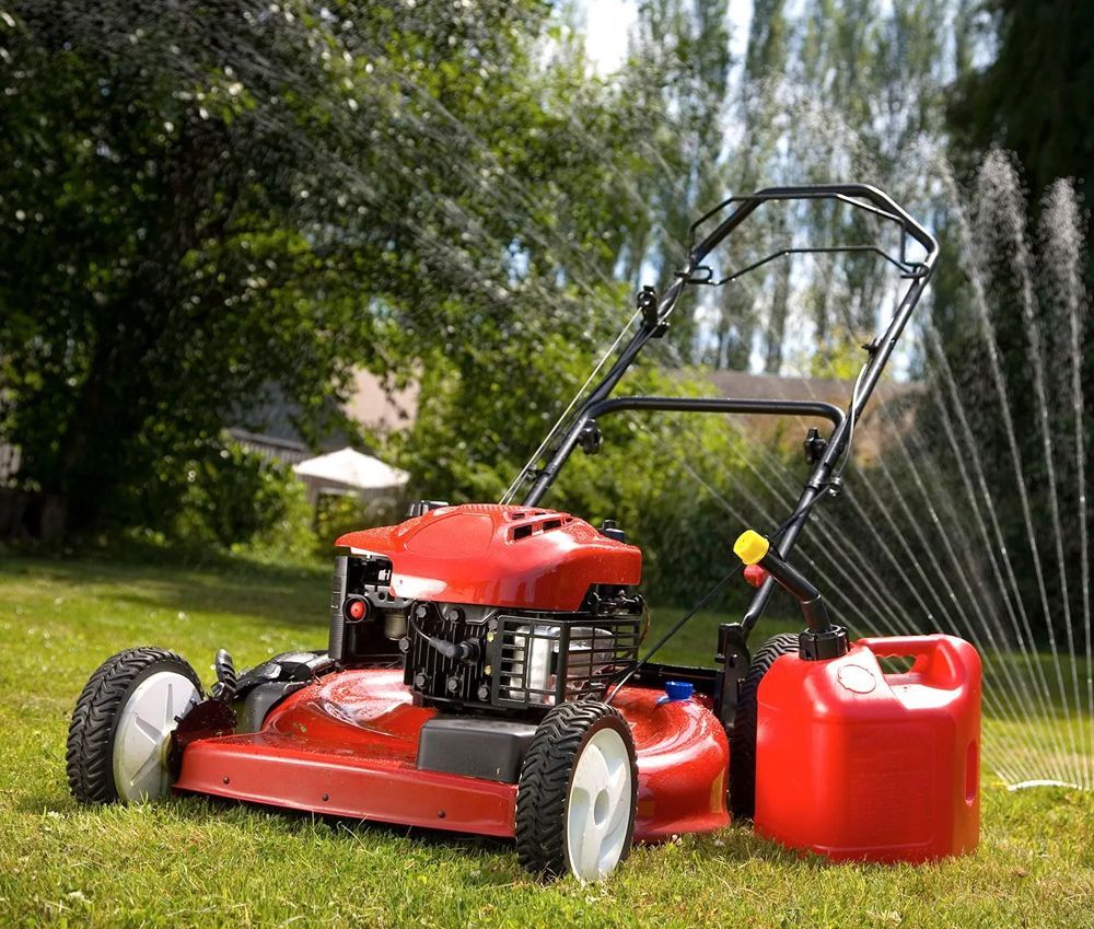 Red lawnmower on grass with a red gas can; water sprinkler in the background.