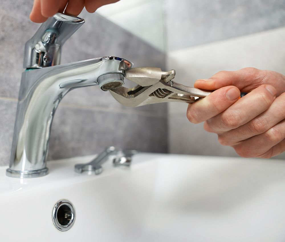 Person using a wrench to repair a chrome faucet in a white sink, in front of a gray tile wall.