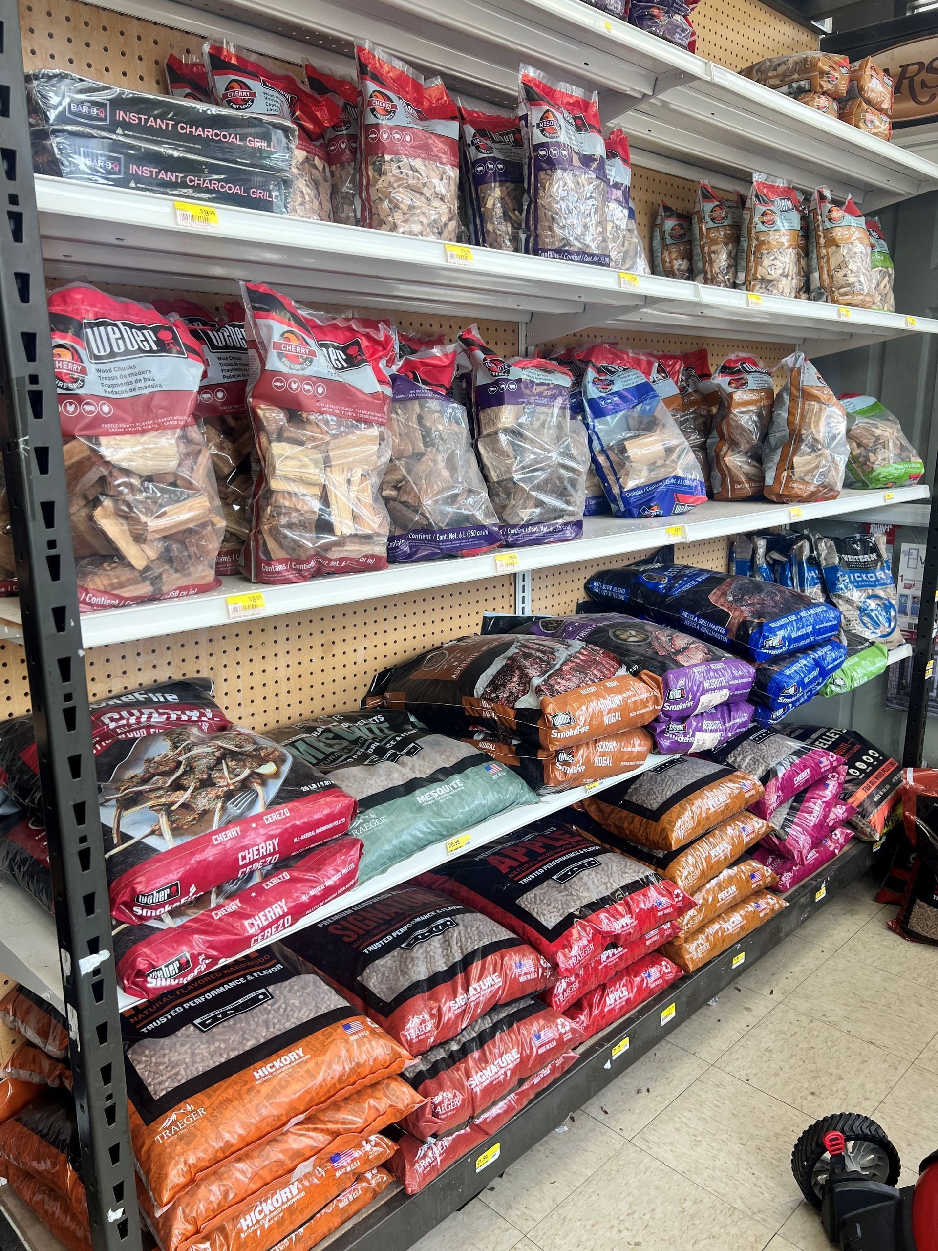 Shelves stocked with various wood chips and pellets for smoking/grilling. Bags in various colors, store setting.