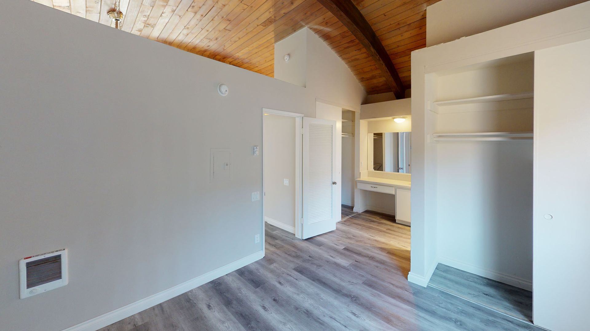 Interior room with vaulted wood beam ceiling, open closet, and bathroom. Grey walls and wood-look flooring.